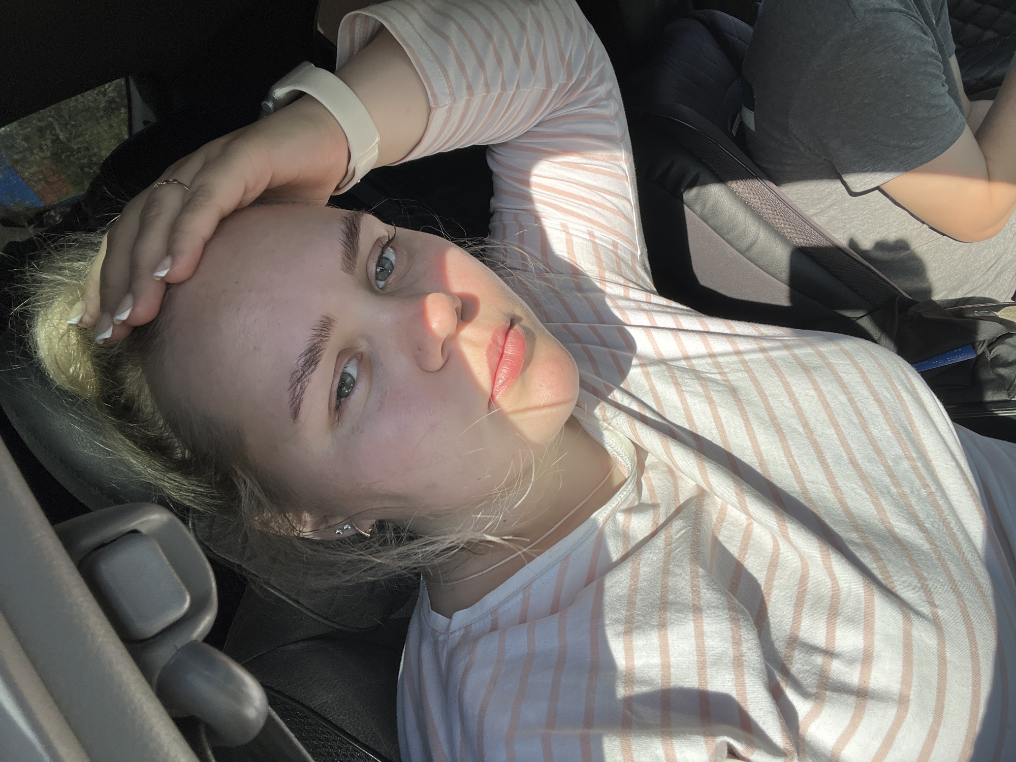 A woman with blonde hair and blue eyes is lying in a car seat. She is wearing a white and pink striped shirt and a white smartwatch on her left wrist. Her hand is resting on her forehead. The car interior is visible, including the seat and seatbelt. The location is likely Chaikovsky, Russia.
