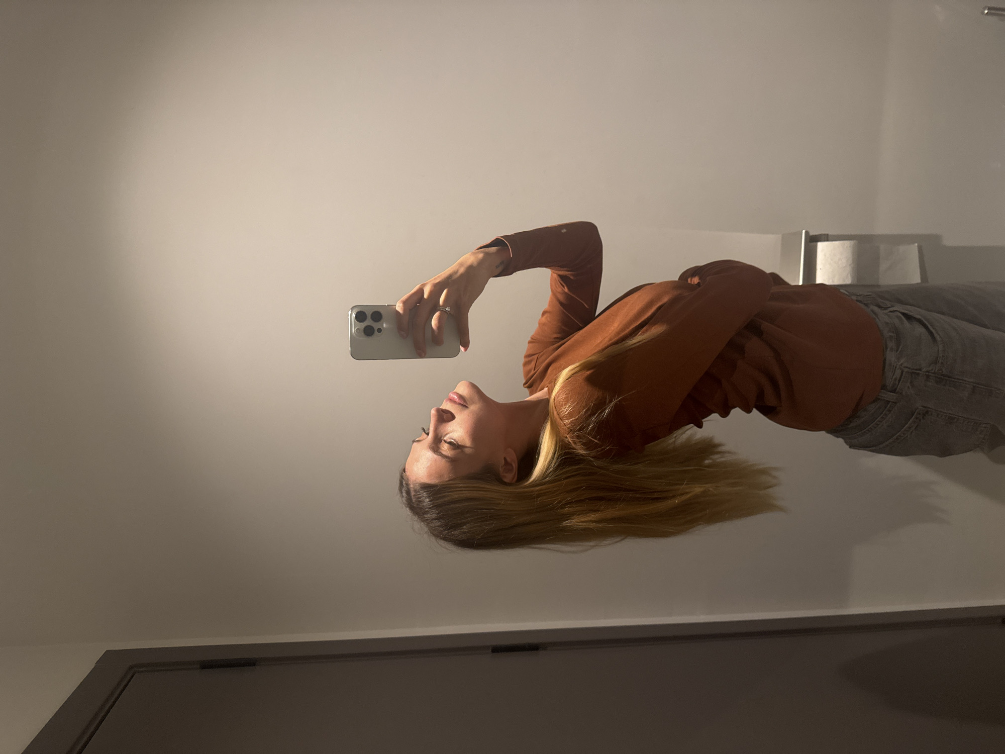 A woman with long blonde hair is holding a smartphone in her right hand and looking at it. She is wearing a brown long-sleeved shirt and light-colored jeans. The background is a light-colored wall. A roll of toilet paper can be seen in the background, slightly behind her. The lower portion of a door is also visible along the bottom of the image. There is no way to determine that this photo was taken in Tbilisi, Georgia.