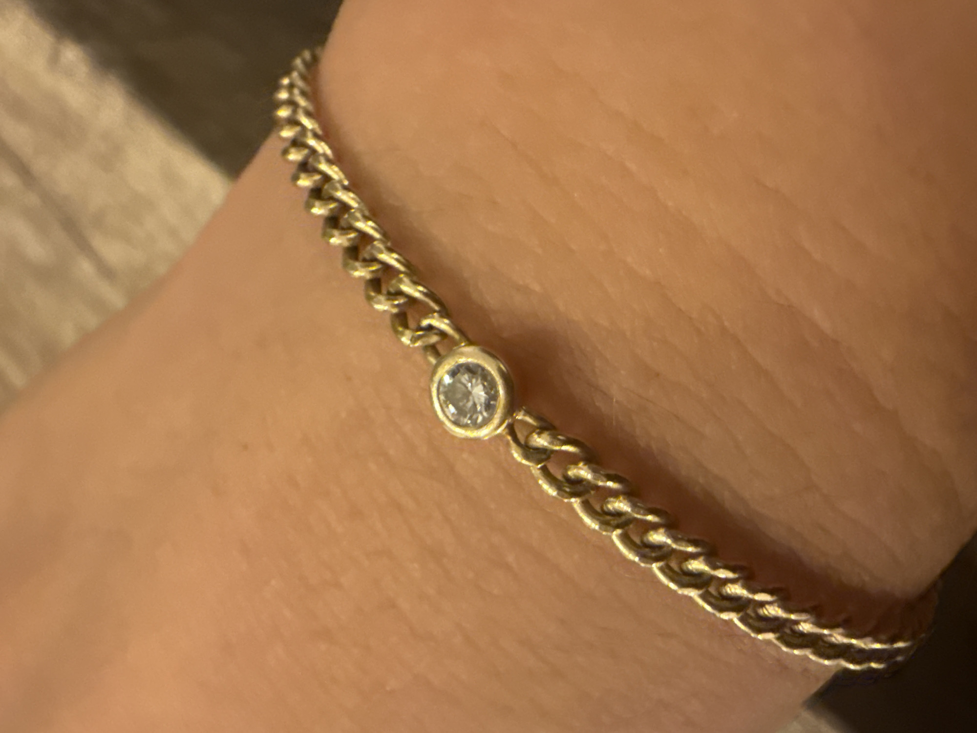 A gold chain bracelet with a single bezel-set diamond is worn on a person's wrist. The bracelet is positioned diagonally across the wrist, with the diamond centered on the upper half of the image. The skin texture is visible beneath the bracelet, indicating the bracelet is on a human arm. The background is blurred and appears to be a textured surface, possibly wood. The image is taken in a close-up, top-down perspective. There is no discernible location in the city of Sozopol, Bulgaria.