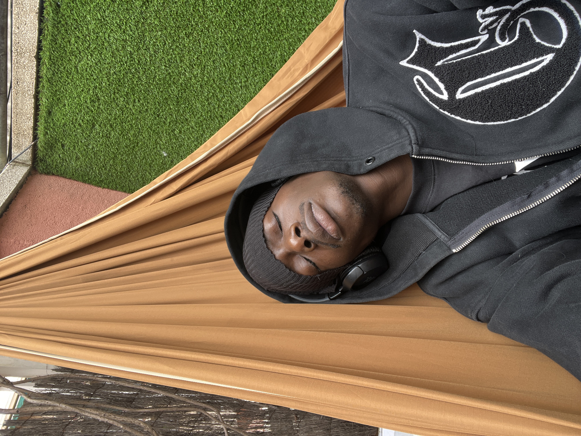 A man is lying in a hammock on a balcony in Barcelona, Spain. He is wearing a black hoodie with a white logo, a black beanie, and headphones. The hammock is tan, and there is artificial grass on the balcony floor. The hammock is hung near some branches.