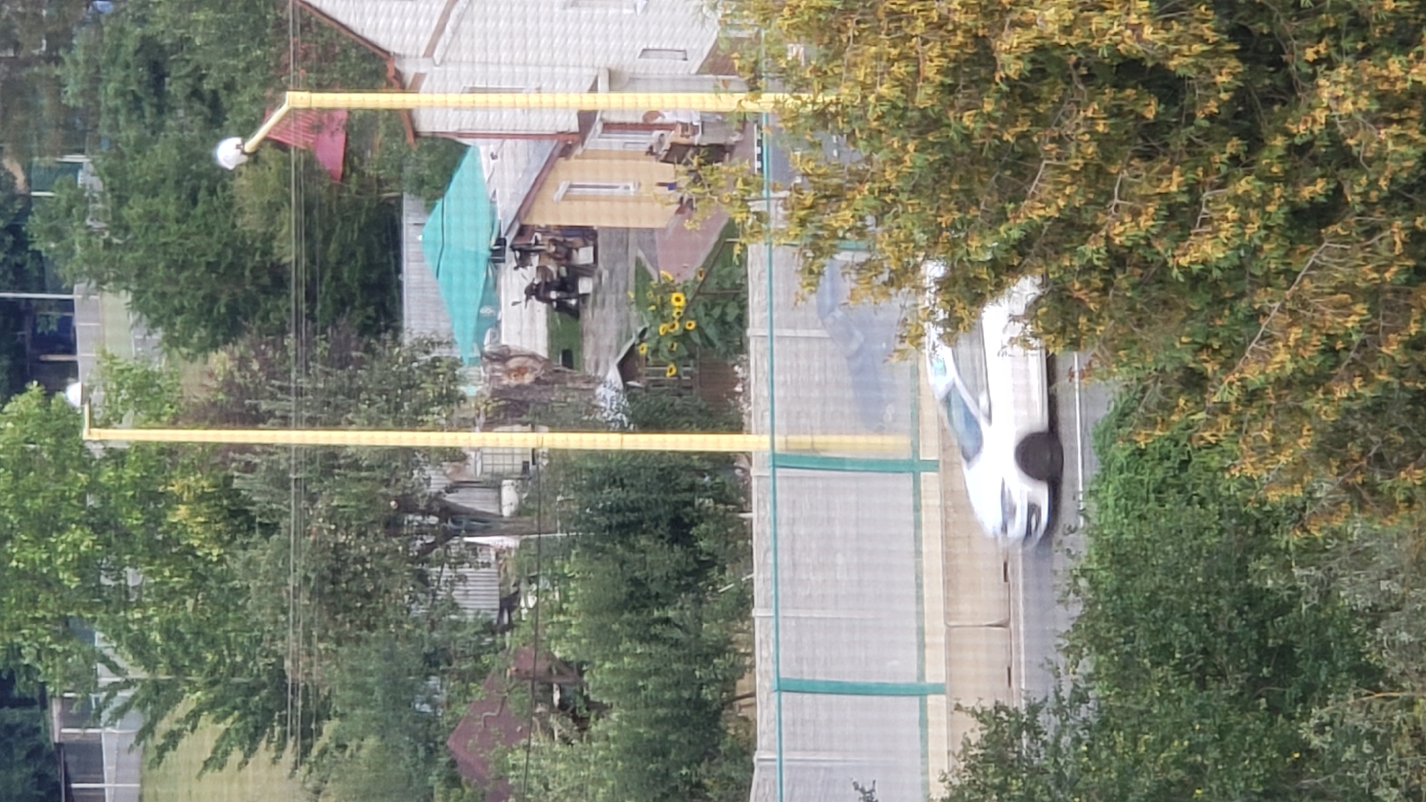The image captures a view from an elevated perspective overlooking a road with a white car driving on it. The road is bordered by a sound barrier wall made of concrete segments with a green mesh overlay. Beyond the barrier, a residential area is visible, featuring houses, a small shed, a turquoise umbrella, and several trees with green and yellow foliage. Two tall yellow poles with lights are prominent in the scene. In the foreground, a tree with yellowing leaves partially obscures the view. The overall setting suggests a suburban or semi-urban environment on a clear day.