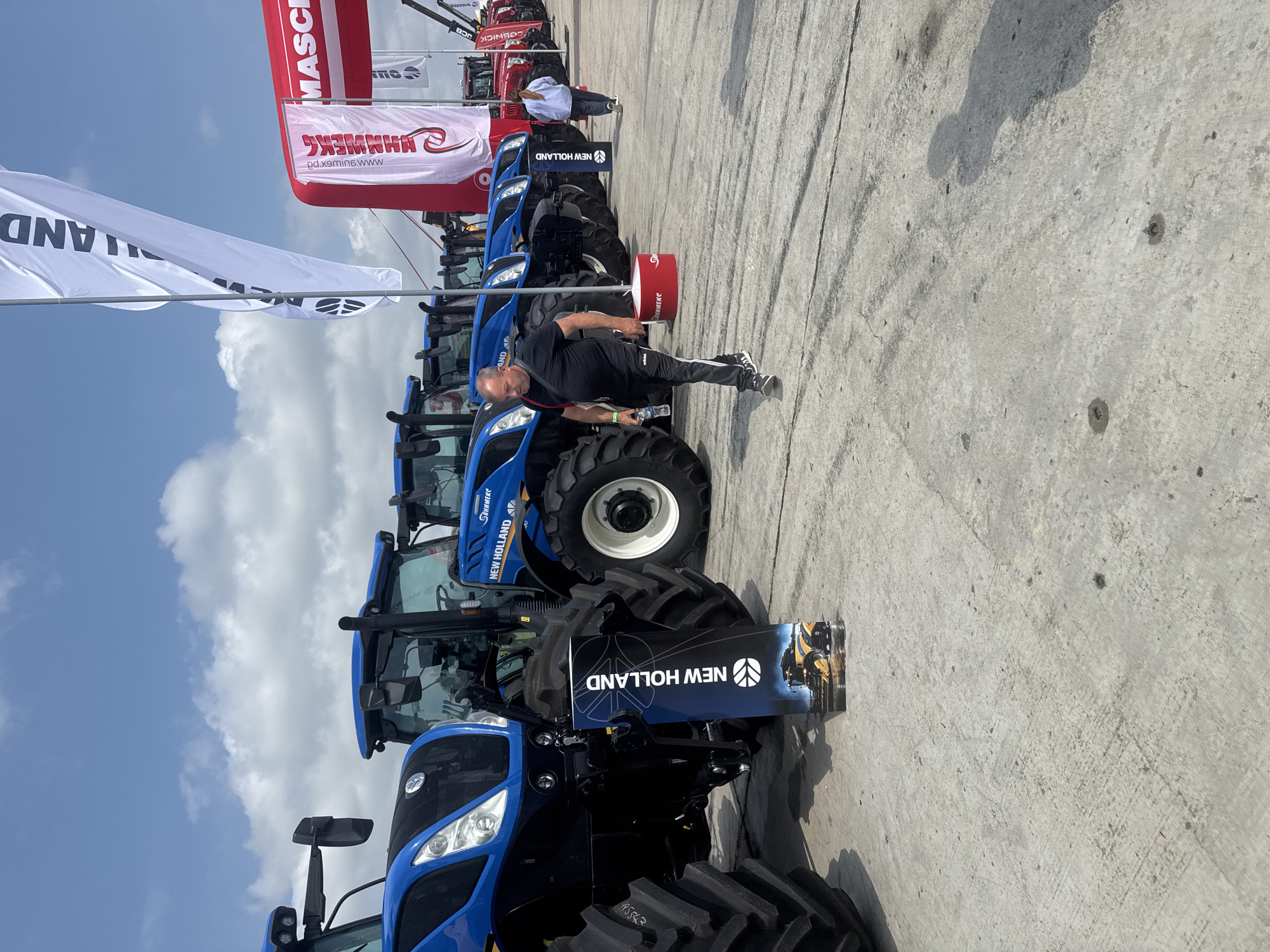 An outdoor scene captures an agricultural machinery exhibition or dealership event in Stara Zagora, Bulgaria, under bright daylight with a clear sky and scattered clouds. The foreground and midground feature a line of several large, blue New Holland agricultural tractors parked on concrete pavement.
A man, dressed in a black t-shirt, black pants with white stripes, and black and white sneakers, stands next to one of the blue tractors in the midground, holding a small bottle. His back is partially to the camera, and he appears to be observing the surroundings or taking a break.
In the background, various promotional flags and banners are visible. A large white flag with black text, partially reading "NEW HOLLAND" and "AND", is prominent on the left. Further back, red and white banners display text such as "ГАНИМЕКС" (GANIMEX), "HAMMEX", and "MASC", with a website "pd.x9mins.www" also visible. Other vehicles, possibly construction equipment or more agricultural machinery from brands like "COBNICK JCB", can be seen further in the distance. A red cylindrical object, possibly a waste bin or small display, is positioned near the tractors. One of the tractor tires has the number "985543" printed on it. The scene conveys a commercial or informational gathering focused on agricultural equipment.