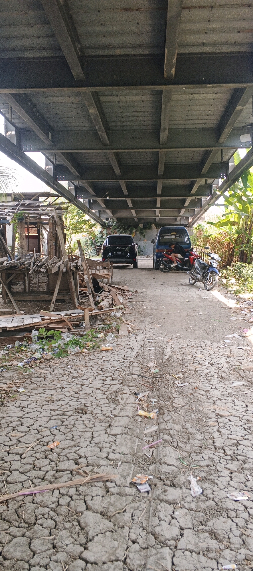 An outdoor shot captures a perspective looking forward from underneath a large, industrial-style metal structure, possibly a bridge or overpass, in the Laren area of Indonesia. The ground is a cracked, dry, dusty surface littered with debris, including plastic bottles, wrappers, and scattered wood fragments. To the left, a makeshift wooden structure is piled high with wood scraps and other discarded materials. Further back, a dark-colored SUV is parked. Next to it, a blue van has a person sitting on the driver's side, seemingly resting. In front of the van, a red motorcycle is parked with a blue motorcycle parked beside it. The background shows lush green foliage and trees. The overall scene suggests an area that is somewhat neglected or under construction, with evidence of human activity and a mix of vehicles and dilapidated structures.