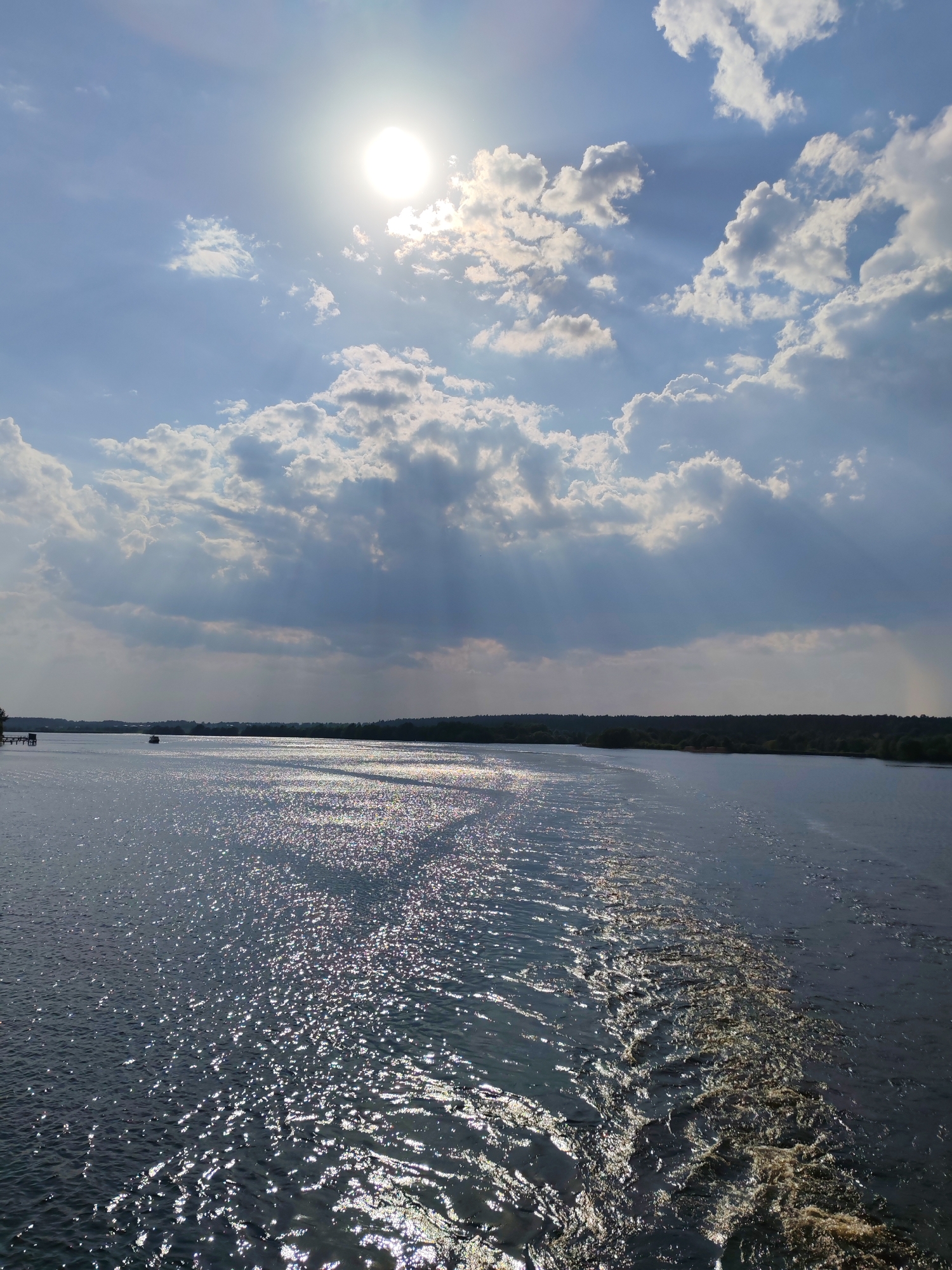 The image presents a wide expanse of water under a bright, partially cloudy sky. The sun is high in the sky, partially obscured by clouds, with sunbeams filtering through them. The water is shimmering with reflected sunlight, with visible wake patterns suggesting movement through the water. In the distance, a dark treeline is visible along the horizon, with a few indistinct structures on the left side of the frame. There is no evidence of location or mention of Nutromo, Russia in the visible content.