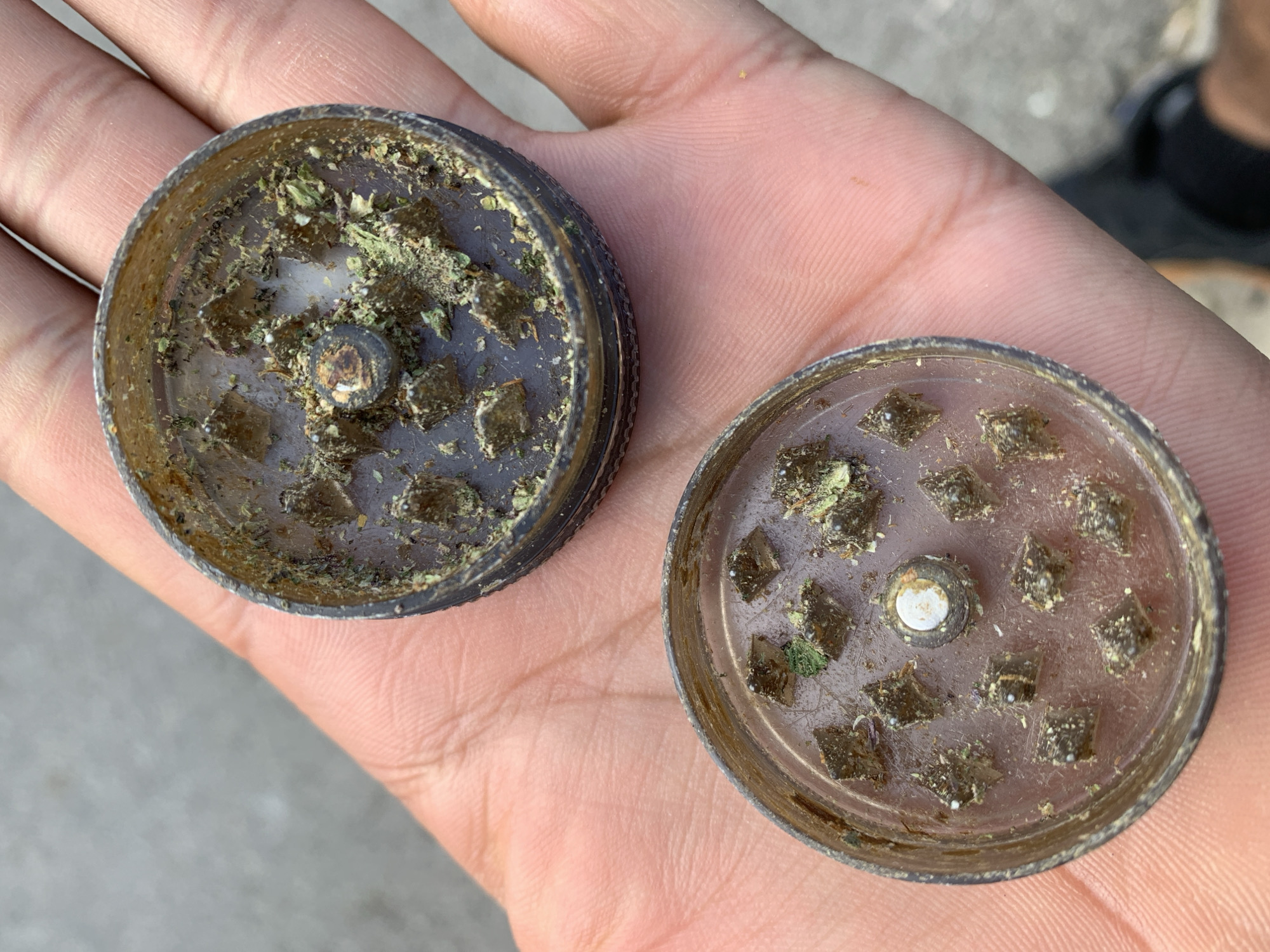 Two disassembled parts of a metal herb grinder are held in a person's open palm, revealing the grinding teeth and remnants of ground herbs within. The background suggests an outdoor urban setting with a blurred grey surface, possibly asphalt or concrete, characteristic of a street or sidewalk. The image is taken from a high angle, looking down into the hand. There are no discernible landmarks to pinpoint a specific location within Barcelona, Spain.
