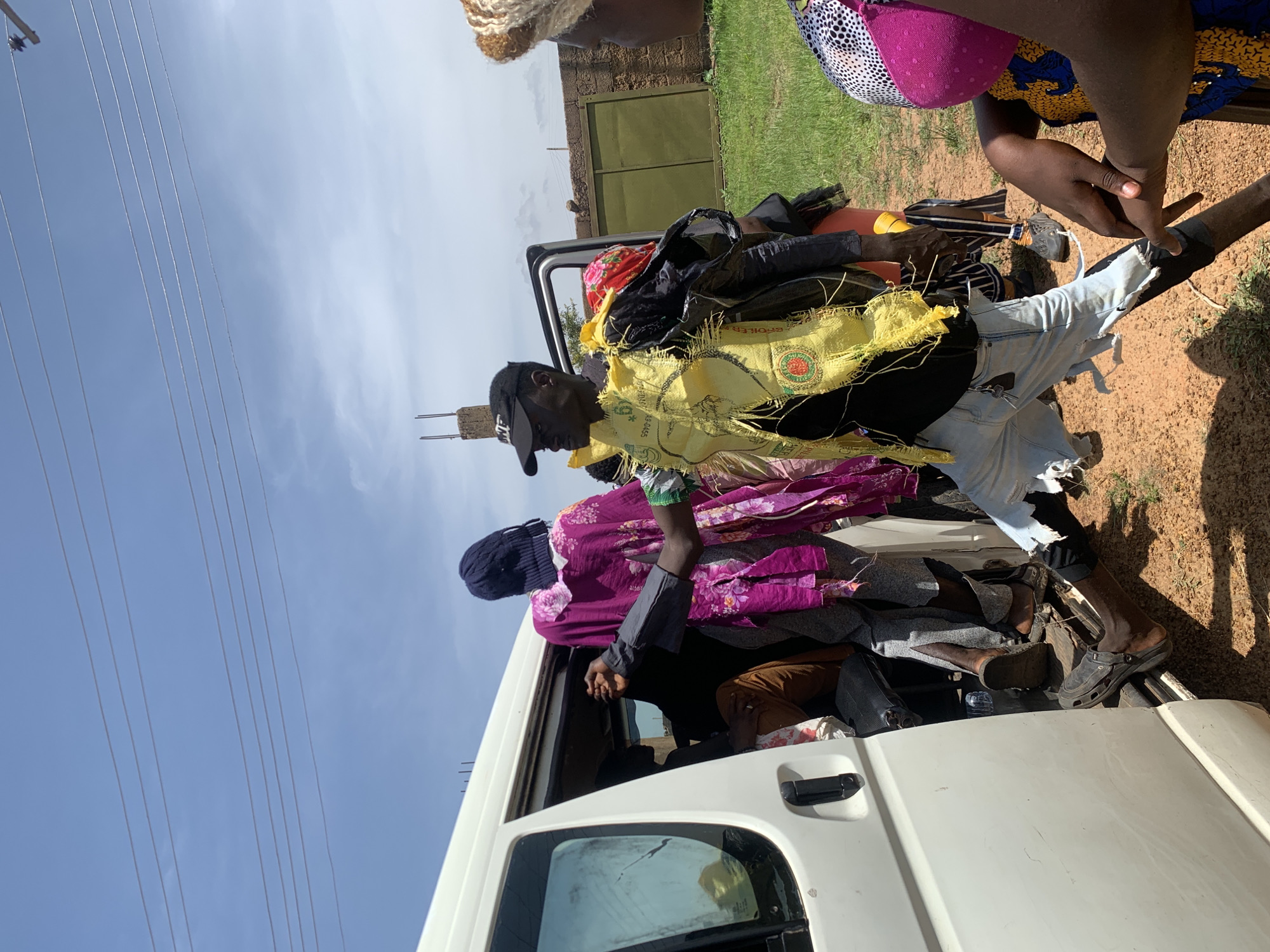 In Gwafan, Nigeria, a group of people are gathered around the open door of a white van. A man in the foreground, wearing a black baseball cap and a torn yellow vest over a black shirt, stands with his left leg extended. He has a red and black patterned cloth wrapped around his head and what appears to be a black bag slung over his shoulder. To his left, a person wearing a vibrant purple garment with a floral pattern and a dark blue head covering is partially visible, reaching into the van. Inside the van, several individuals are seated or standing, with one person in brown pants and another in grey pants clearly visible. To the right of the man in the yellow vest, a person wearing a colorful patterned top and a pink head covering is seen from the side, with their hands clasped in front of them. The ground is covered in reddish-brown dirt and patches of green grass. In the background, a low brick wall with a green gate is visible, and power lines stretch across the bright blue sky with scattered clouds. A partially constructed concrete pillar is also present in the background.