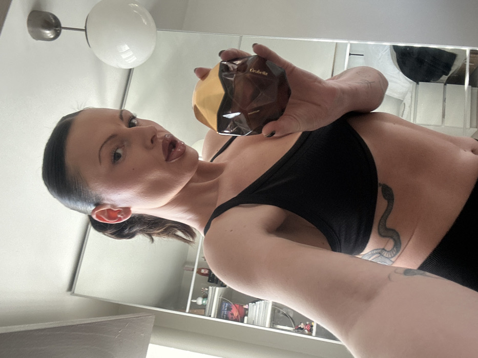 A female individual is depicted indoors, visible from the mid-torso up, holding a perfume bottle. Her dark hair is styled in a ponytail. She wears a black ribbed bralette, revealing a dark snake tattoo on her left side, below the ribcage. Her facial expression features parted lips with a partially visible tongue, while she looks directly at the viewer. She holds a faceted, dark brown 'Orabella' perfume bottle with a gold-colored, irregular top in her right hand. Her fingernails are painted dark. The background is a reflection in a large mirror or mirrored wardrobe, revealing a white ceiling with a spherical pendant light fixture. A shelving unit containing books and various personal items is also visible in the reflection. Further into the reflected background, white furniture and a dark garment are discernible. The precise location within the city of Wealden, United Kingdom, cannot be determined from the provided image.