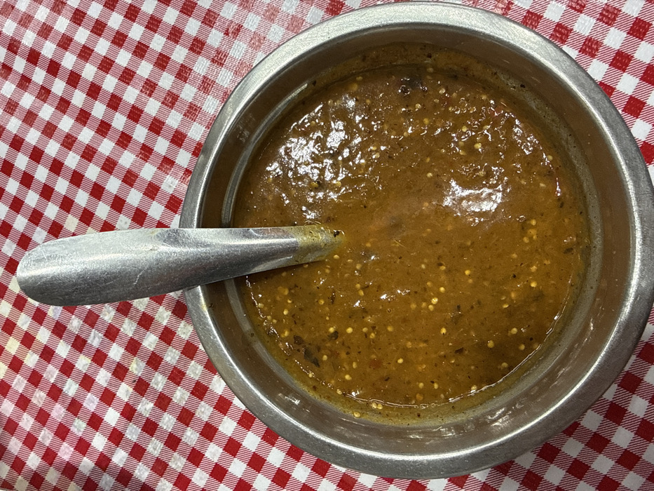 A close-up, overhead view depicts a metal bowl containing an amber-brown salsa with visible seeds and specks of darker ingredients. A silver-colored metal spoon is partially submerged vertically within the salsa, its handle extending upwards and slightly to the left. The bowl is positioned on a surface covered by a red and white checkered (gingham) tablecloth. The scene is located in the city of Nezahualcóyotl, Mexico.