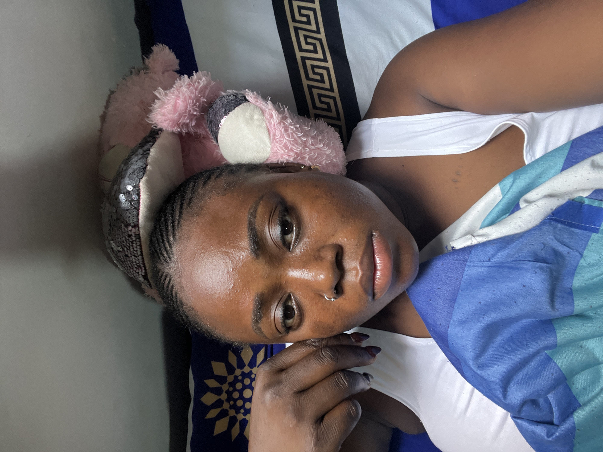 The image is a close-up selfie taken indoors, likely in a bedroom, in Abuja, Nigeria. The subject is a young Black woman lying down, with a pink stuffed animal and a sequined item partially visible behind her head. She is wearing a white tank top and a blue and white striped blanket is covering her. Her face is the primary focus, with her hand resting on her cheek. The background consists of pillows and bedding with patterned designs. The lighting suggests it is daytime, possibly with natural light filtering in.