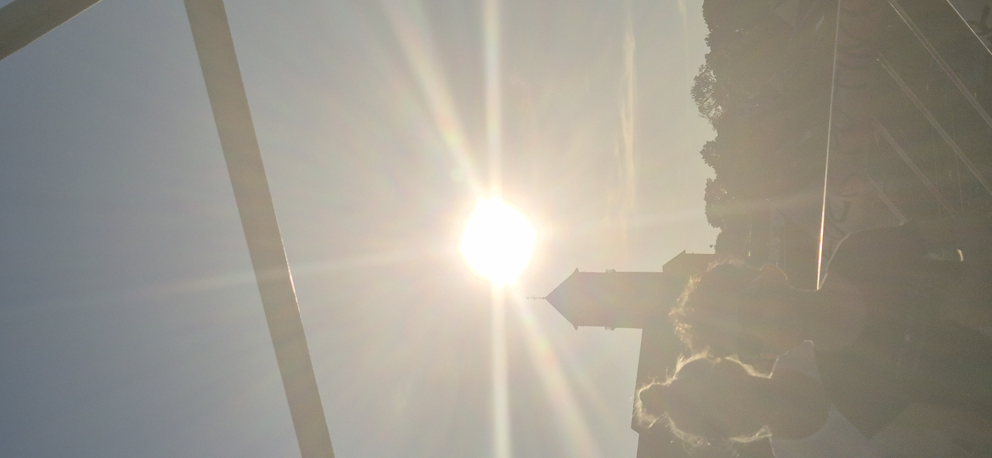 The image depicts a bright, sunny day in Mainz, Germany, with the sun positioned high in the sky, casting a strong glare. In the foreground, the backs of two individuals are visible, their faces obscured by the bright light. They appear to be standing or sitting, looking out towards a cityscape. Behind them, a prominent tower, likely part of a church or historical building, is silhouetted against the sky. Lush green trees form a dense backdrop to the scene. The overall atmosphere is one of a pleasant, possibly tourist-oriented, outdoor setting in a German city.