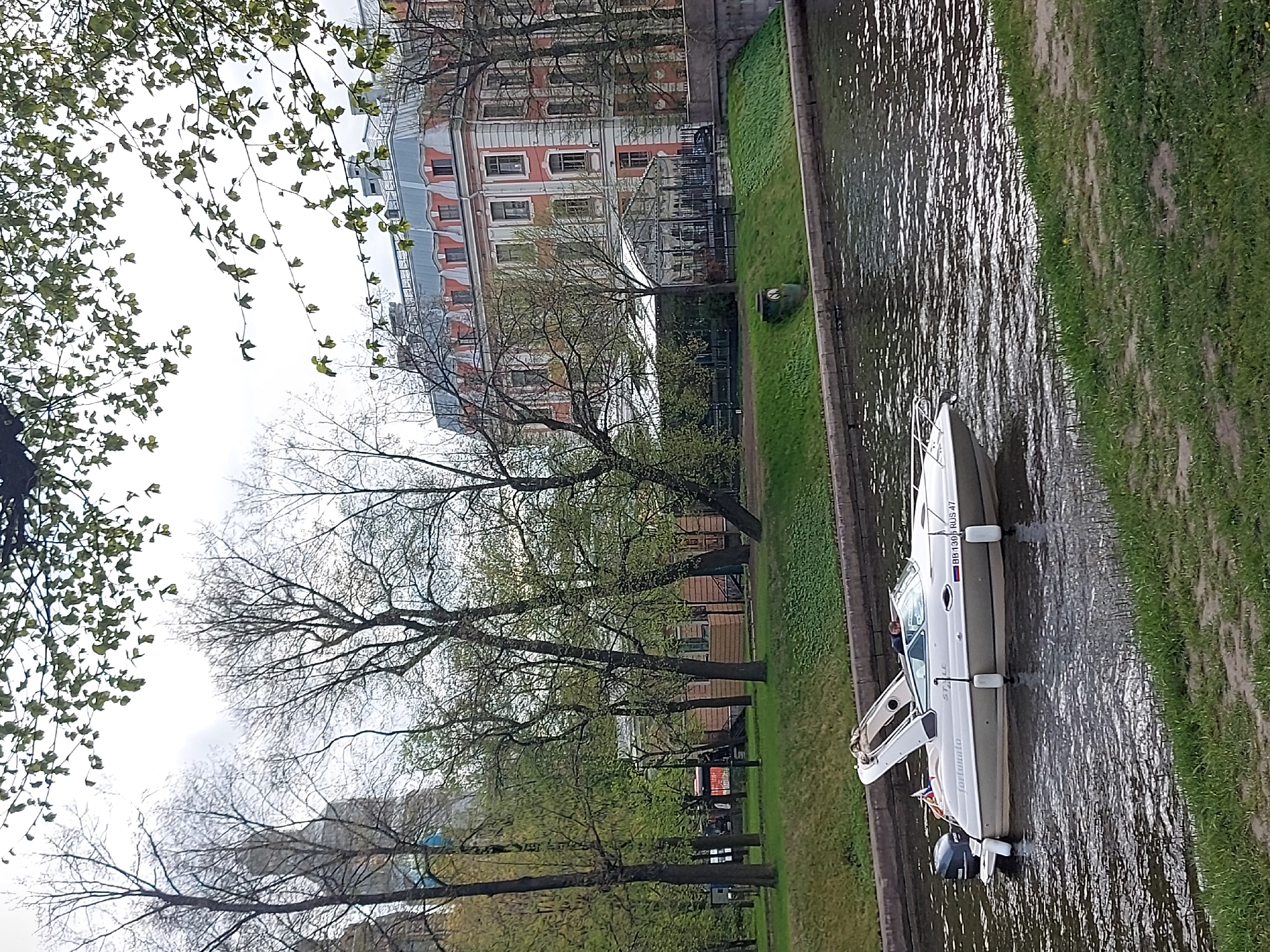 The image captures a canal in Saint Petersburg, Russia, with a white motorboat "Fontanalo" moored along the concrete embankment. The boat bears the registration number "BB 1395 RUS-47." Lush green grass lines the canal's edge, creating a striking contrast with the water. Across the canal, a line of trees partially obscures the view of buildings with a reddish facade and a grey roof. The trees are a mix of bare branches and newly sprouting leaves. The sky appears overcast.