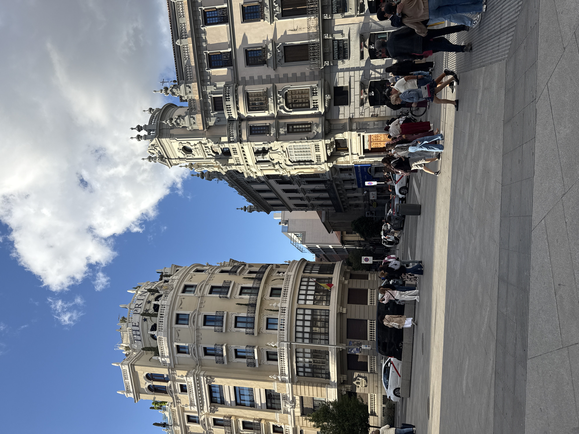 The image depicts a street scene in Madrid, Spain, with two ornate buildings framing the view. The building on the left, partially visible, is labeled "THE PRINCIPE" at the top. A Spanish flag is visible on one of the balconies. The building on the right showcases elaborate architectural details and balconies. Pedestrians are present on the sidewalk, including individuals standing and walking. Some people are wearing masks. The sky is blue with white clouds. Parked cars and taxis line the street. The sidewalk is paved with large gray tiles. The image is taken at a slightly tilted angle.