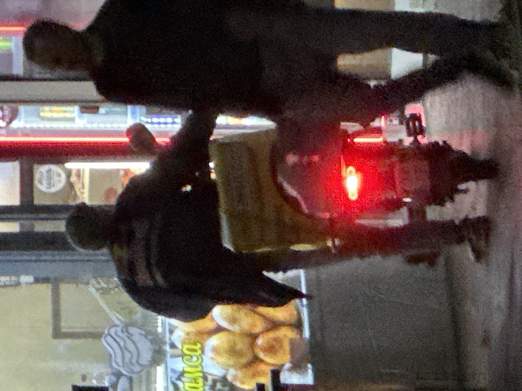 Two individuals are depicted outdoors in Podolsk, Russia, near a food establishment. The person on the left, wearing dark clothing and a helmet, is on a scooter with a large yellow food delivery box. Their right leg is extended, and they appear to be interacting with or gesturing towards the person on the right. The person on the right, also in dark clothing, stands facing away from the camera. Behind them, the storefront displays a variety of food items, possibly pastries or bread, and a refrigerated beverage display with illuminated bottles. The lighting suggests it is nighttime or dusk, with bright neon lights from the establishment illuminating the scene and reflecting on the wet pavement. A red taillight from the scooter is prominently visible.