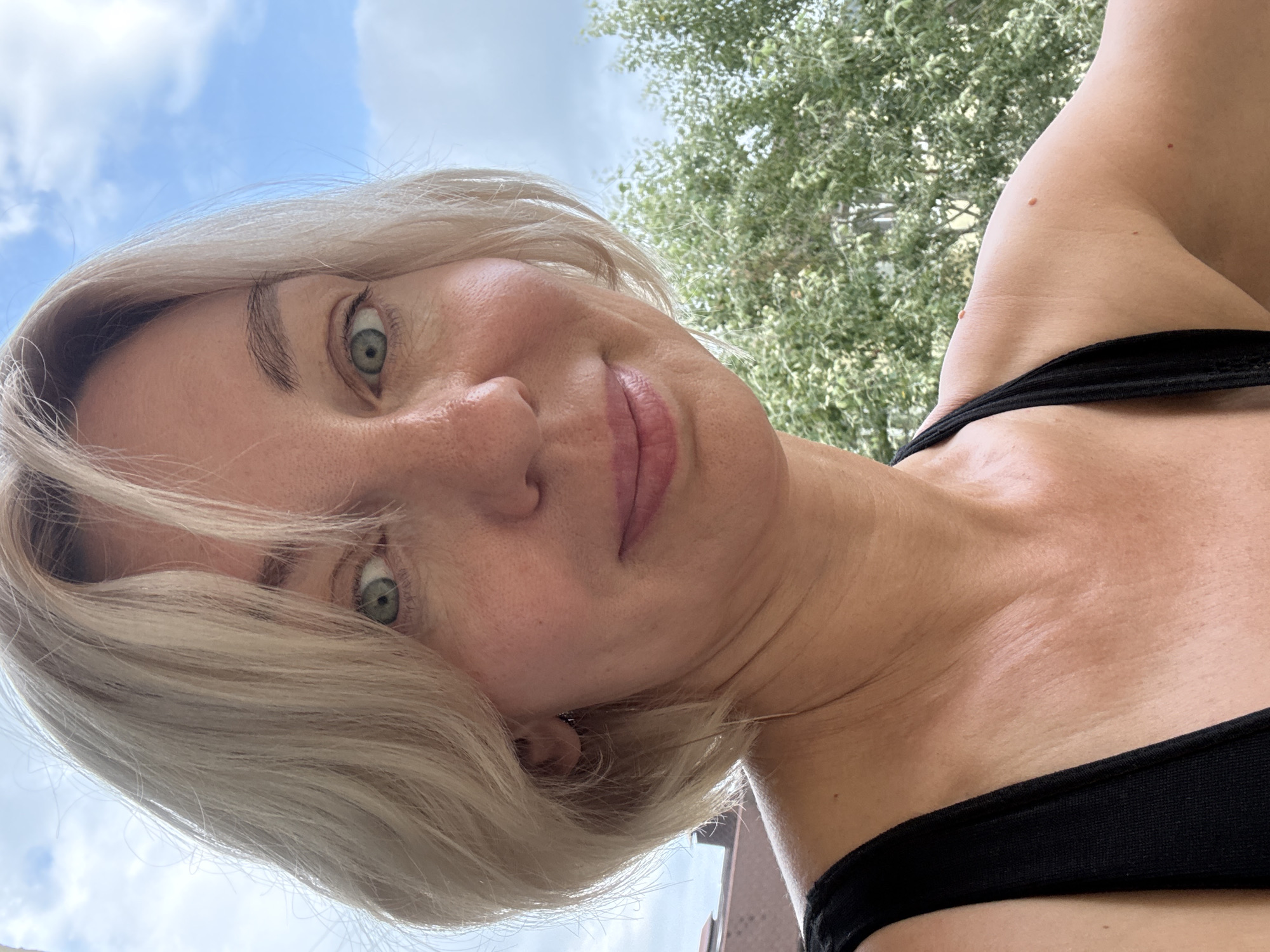 A woman with blonde bob-length hair and green eyes smiles directly at the camera. She is wearing a black tank top. In the background, green foliage and a cloudy blue sky are visible. The scene appears to be outdoors, potentially on a balcony or terrace, with some architectural elements, possibly buildings, visible in the distance.