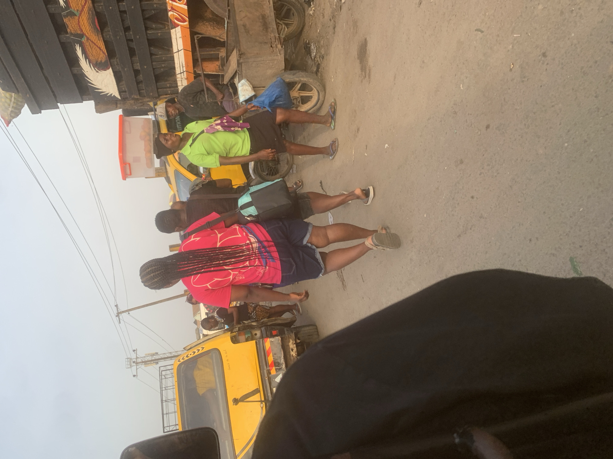The image depicts a bustling street scene in Fegge, Nigeria, under an overcast sky. Multiple individuals are present, engaged in various activities related to daily commerce and transportation.
In the mid-ground, several people are visible. One person, seen from behind, wears a bright pink shirt with a white pattern and dark shorts, carrying a red backpack. Another individual in a lime-green shirt and dark shorts stands near a yellow three-wheeled vehicle, possibly a "keke napep." This person appears to be a vendor or customer, as a red-lidded cooler containing what looks like oranges or similar produce is placed nearby. The cooler has partially visible text, "UTION," on its side.
A large, heavy-duty truck with a distinctive side panel decorated with white feather-like motifs on a dark and orange background is positioned further back. Another person is seen sitting or resting near the truck's wheels. The street surface is a mix of asphalt and dirt, showing signs of heavy use.
Overhead, power lines are visible against the hazy, overcast sky. The yellow vehicle in the background has some faint markings and text on its rear, including what appears to be "Exod 13 v 201." People are dressed casually, with many wearing flip-flops. The scene captures an ordinary daytime street environment, likely a market or a transport hub. No specific strong emotions are evident.