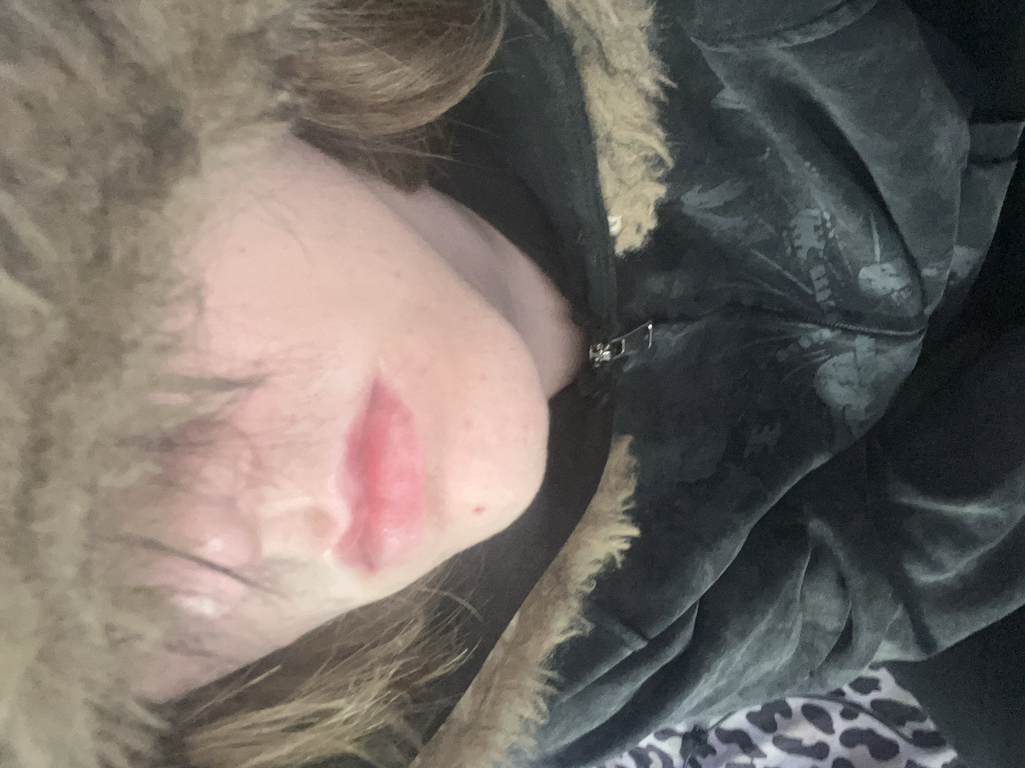 A close-up, angled photograph captures a portion of an individual resting, likely indoors in Saint Petersburg, Russia. The subject's head is tilted, showing their light brown, wavy hair framing the side of their face. Visible facial features include a cheek, chin, and lips, which are slightly parted and appear reddish-pink. The skin tone is fair, with a small red mark visible below the mouth.
The person is wearing a dark-colored, possibly velvet or plush, hooded garment. The hood features a light brown or tan faux fur trim around the edge. A silver-toned zipper is visible on the garment's front. The dark fabric of the garment has a subtle, lighter-toned pattern, possibly depicting leaves or a similar organic motif. In the lower right corner, a glimpse of a grey and white leopard-print fabric, likely a blanket or another piece of clothing, suggests a cozy, relaxed setting. The overall impression is one of rest or relaxation. No text is visible in the image.