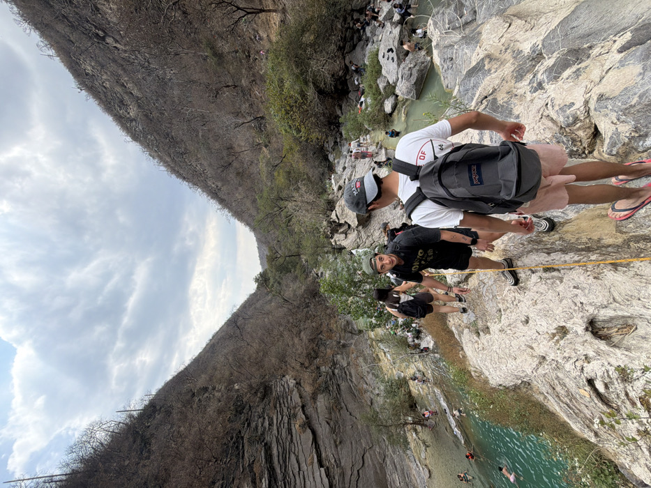 The image displays a natural canyon or valley environment near San Miguel Atlapulco, Mexico, characterized by steep, sparsely vegetated hillsides and a clear, light blue-green river flowing through the bottom. In the immediate foreground, three individuals are positioned on a large, light-colored rocky outcrop, appearing to descend towards the river below. A male on the right, viewed from behind, wears a white t-shirt, pink shorts, a grey baseball cap, and pink flip-flops, carrying a grey JanSport backpack. To his left, another male, facing partially left, wears a dark t-shirt, dark shorts, dark athletic shoes, and a dark cap, grasping a yellow rope that extends downwards. Behind him, a female, also viewed from behind, wears a dark top and dark shorts, carrying a dark backpack.
The canyon walls are composed of layered grey rock formations on the left and steeply rising hillsides covered in dry, brown vegetation on both sides, suggesting a dry season. The sky is largely overcast with prominent cloud formations. In the midground and background, numerous other individuals are visible along the riverbanks and in the water, some swimming or wading, indicating a recreational area. The overall scene depicts a group navigating a rocky descent into a natural river setting populated by other recreationists.