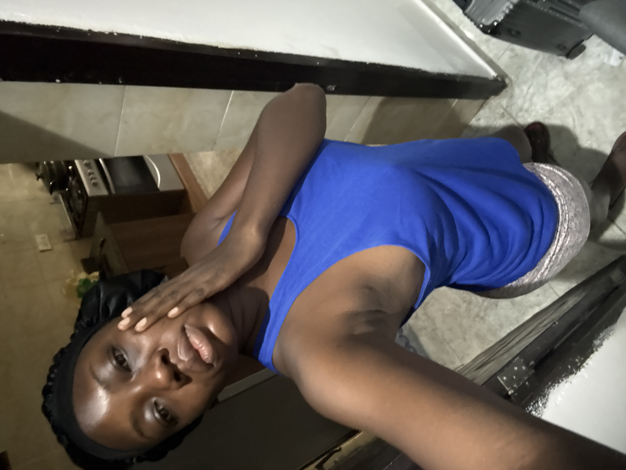 A dark-skinned person is lying on a tiled floor inside a kitchen. They wear a blue tank top, paisley shorts, and a black head covering. Their right hand is positioned near their face. A kitchen counter is visible behind them, with a stove or oven below it. The location is Iddo Tudunwada, Nigeria.