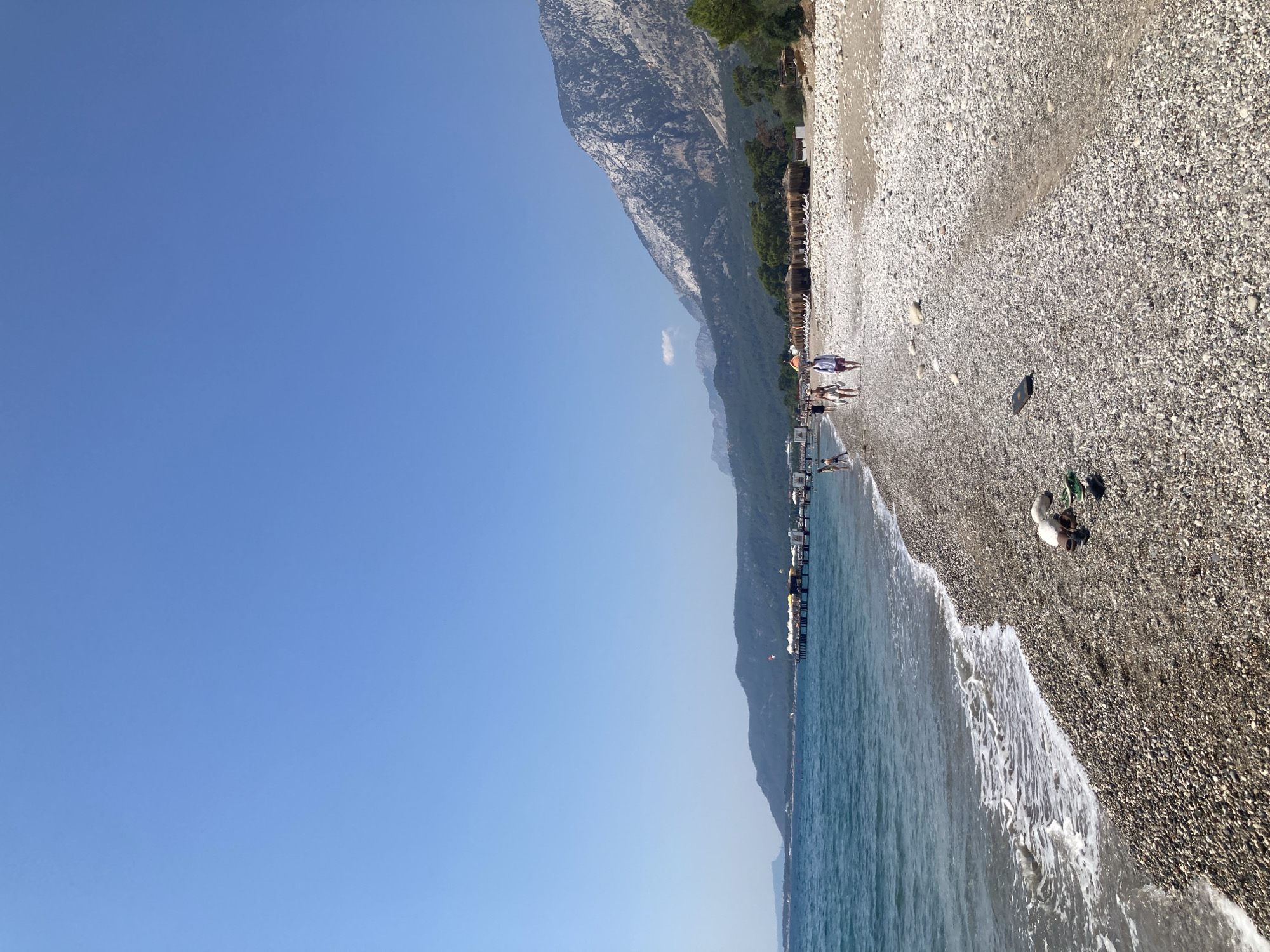 The image captures a scenic view of a pebble beach on a sunny day, likely in Kemer, Turkey. The foreground is dominated by the textured surface of the pebble beach, with small waves gently lapping at the shore. A pair of sandals and some green flip-flops are visible near the water's edge. In the mid-ground, several people are walking along the beach, some wading in the turquoise water, while others stroll on the pebbles. A pier extends into the sea, lined with white umbrellas and lounge chairs, suggesting a resort area. Beyond the pier, a mountainous landscape forms the backdrop, with lush green trees covering the lower slopes and rocky, barren peaks rising towards the clear blue sky. The overall impression is one of a tranquil and picturesque coastal environment.