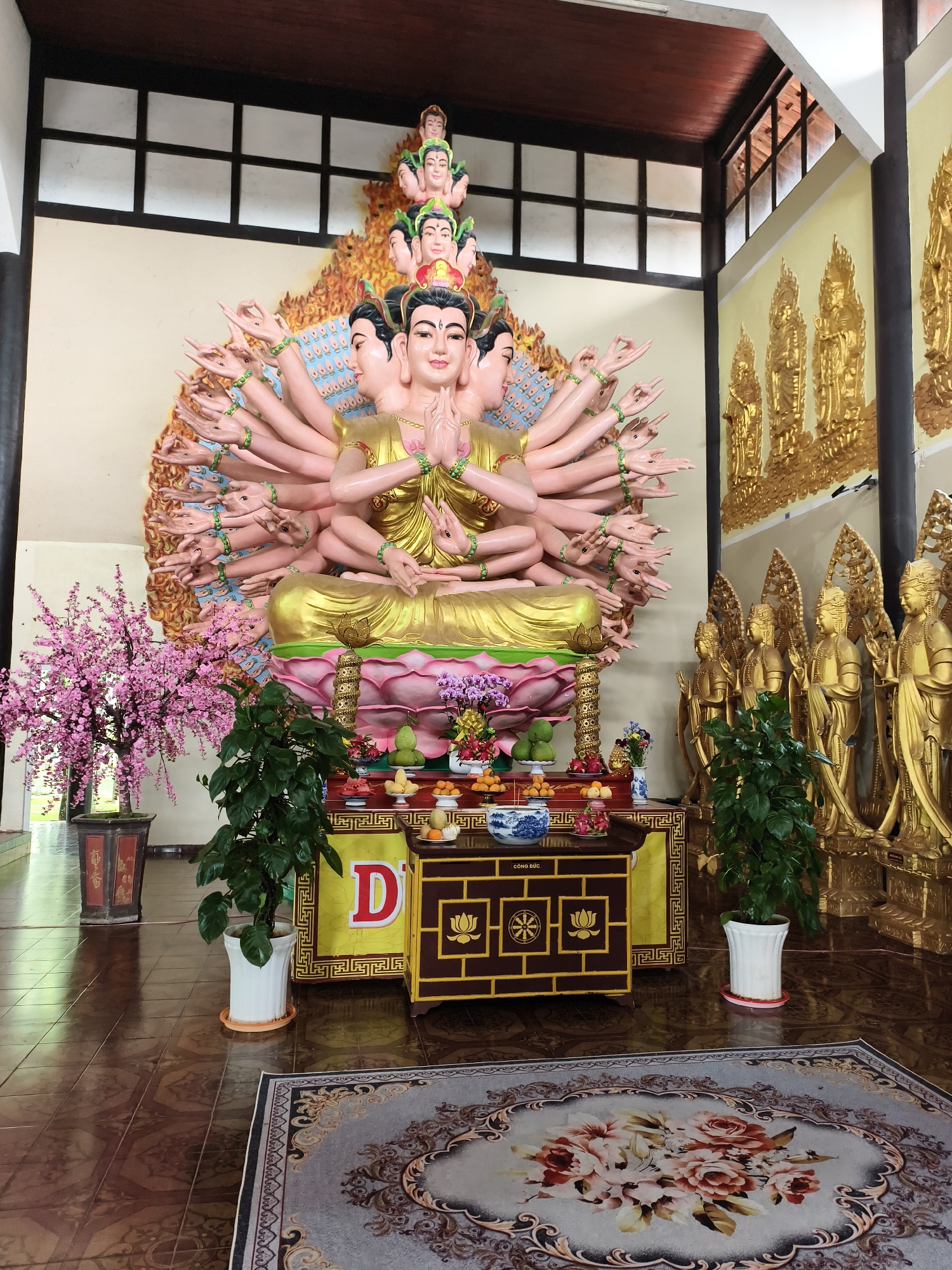 The image shows a large, ornate statue of a Buddha with multiple arms and heads in what appears to be a temple or shrine. The statue is gold and pink, with many arms extending outward. The background behind the statue is also ornate and decorated with golden patterns. In front of the statue is a table adorned with offerings, including fruit and flowers. Potted plants flank the table. A rug with a floral design is on the floor in front of the table. The location is an unknown city and country.