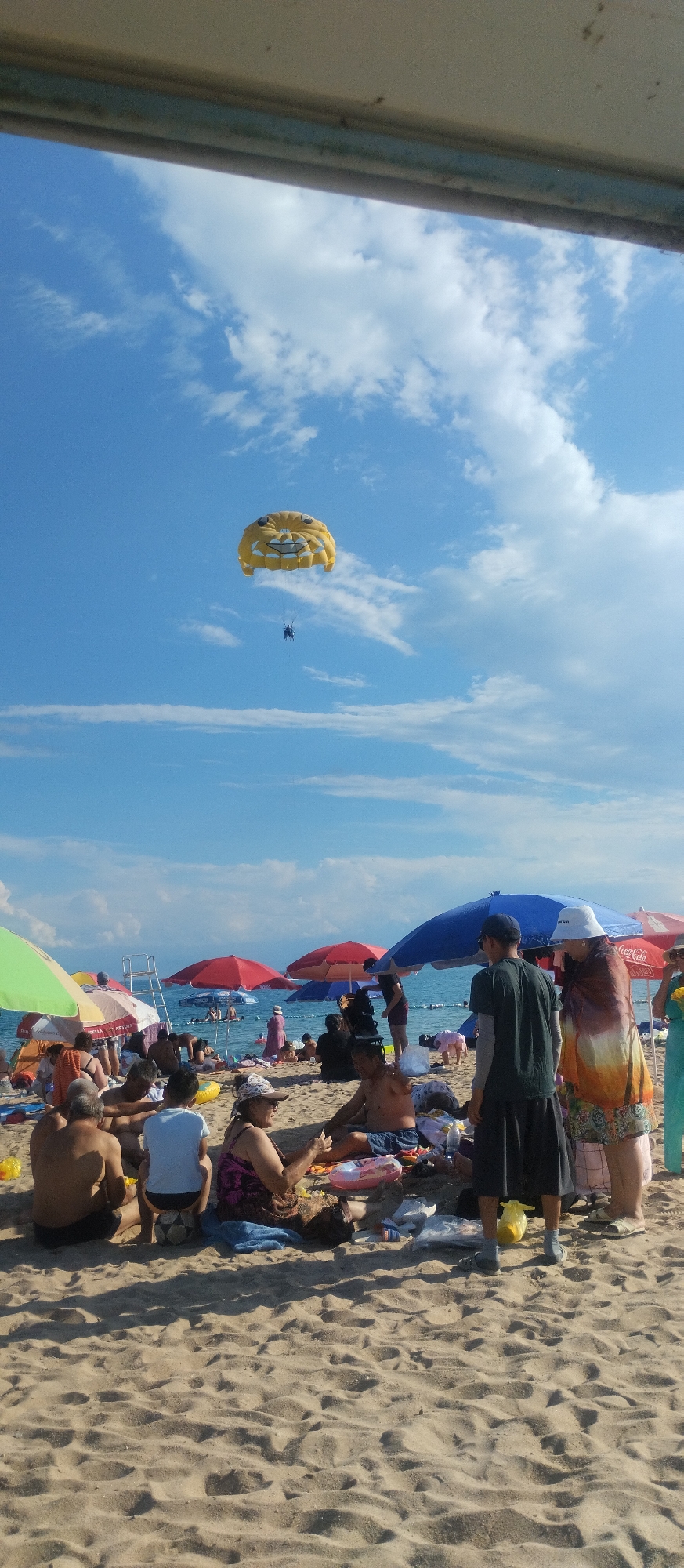 On a sandy beach in Cholpon-Ata, Kyrgyzstan, people are enjoying a sunny day. In the sky, a yellow parasail with a cartoonish face design is visible, with two individuals suspended beneath it. The beach is dotted with various beach umbrellas, including red, blue, and green ones, providing shade for beachgoers. Several groups of people are gathered on the sand, some sitting, some lying down, and some appearing to be interacting with each other. Beach towels, bags, and inflatable rings are scattered around the groups. In the background, the clear blue water of Issyk-Kul Lake is visible, with a few people in the water. The sky is a vibrant blue with scattered white clouds.