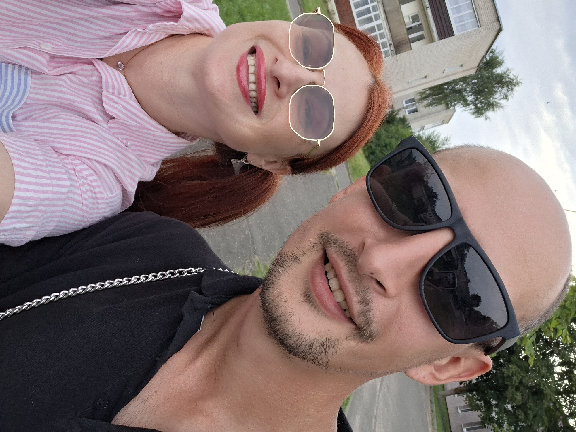 A close-up, selfie-style photograph captures a smiling woman with red hair and a man with a shaved head, both wearing sunglasses. The woman, on the left, sports a pink and white striped shirt and hexagonal-shaped glasses. The man, on the right, is wearing a black polo shirt and a silver chain. In the background, buildings and trees are visible, suggesting an outdoor urban setting. The reflection in the man's sunglasses shows another person holding a phone, presumably taking the photo. The architectural style of the buildings, characterized by beige brickwork and balconies, is consistent with the Soviet-era apartment blocks found in Myadzyel, Belarus. The overall scene depicts a casual moment between two individuals in a residential area.