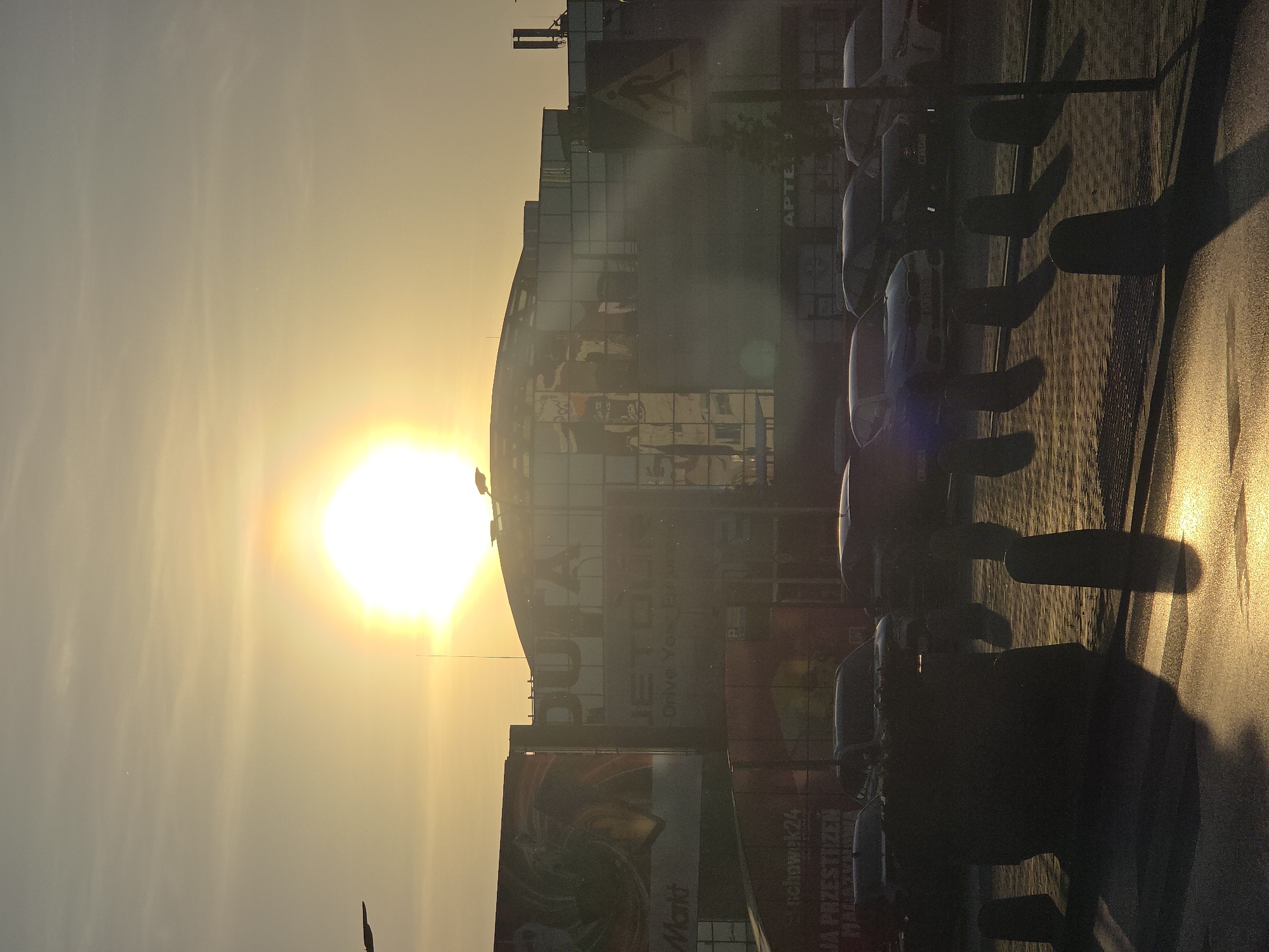 The image captures a cityscape at sunset in Warsaw, Poland. A large, partially mirrored building dominates the view, with various advertisements and signage visible on its facade. "APTEKA" is prominently displayed. In the foreground, a row of parked cars casts long, dark shadows on the street due to the low angle of the sun. The sun, a bright orb, is positioned behind the building, casting a golden glow over the scene.