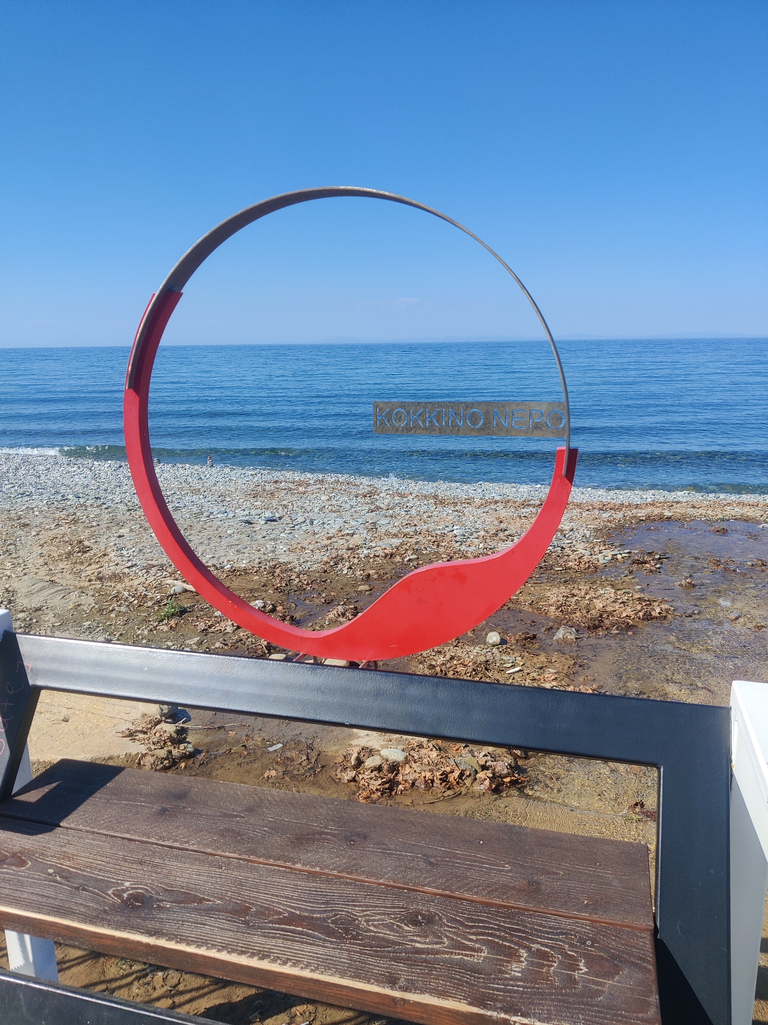 A red and silver circular sculpture is positioned on a beach. The sculpture has text that reads "KOKKINO NERO". The beach has gray pebbles and some dried leaves scattered on it. The sea is calm and blue, with a clear sky above. A wooden bench with a black metal frame is in the foreground. This location is identified as Kokkino Nero, within the Municipal Unit of Evrymenon, Greece.