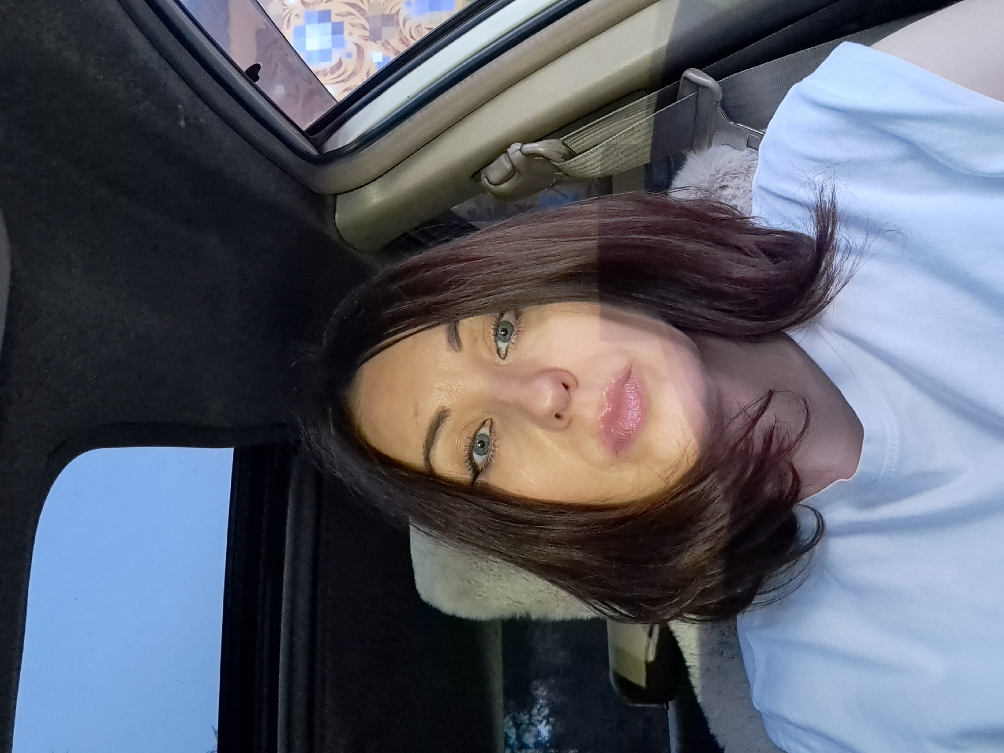 The image shows a woman sitting in a car, with a sunroof open above her. She is looking directly at the camera, with her mouth slightly open and her lips glossy. She has dark brown hair, which is styled in a bob, and she is wearing a white t-shirt. The car's interior is visible, including a seatbelt and part of the car door. The background shows a blue sky through the sunroof, with some greenery visible outside the car. The image was taken in Irkutsk, Russia.