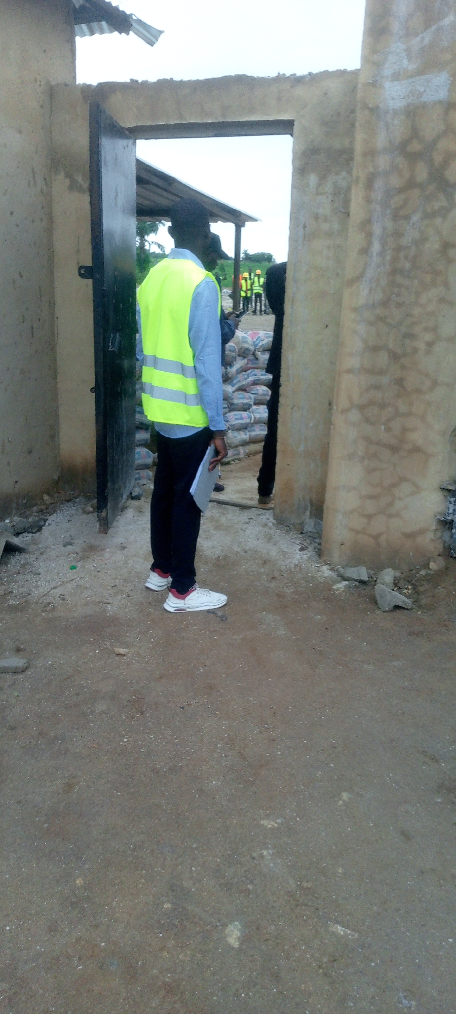 A person wearing a bright yellow safety vest over a blue long-sleeved shirt and dark pants stands facing away from the camera. They are holding a paper document. White sneakers with red accents are on their feet. They are standing in what appears to be a construction or material storage area, with bags of cement stacked in the background. The setting seems to be outdoors, with an overcast sky visible. To the left, a dark metal door is ajar, part of a rudimentary building made of rough, light-colored material. In the distance, through an opening, other individuals in similar safety vests and hard hats can be seen, suggesting a work site. The ground is dusty and unpaved, with scattered stones and debris. The time of day appears to be daytime, under cloudy weather conditions.