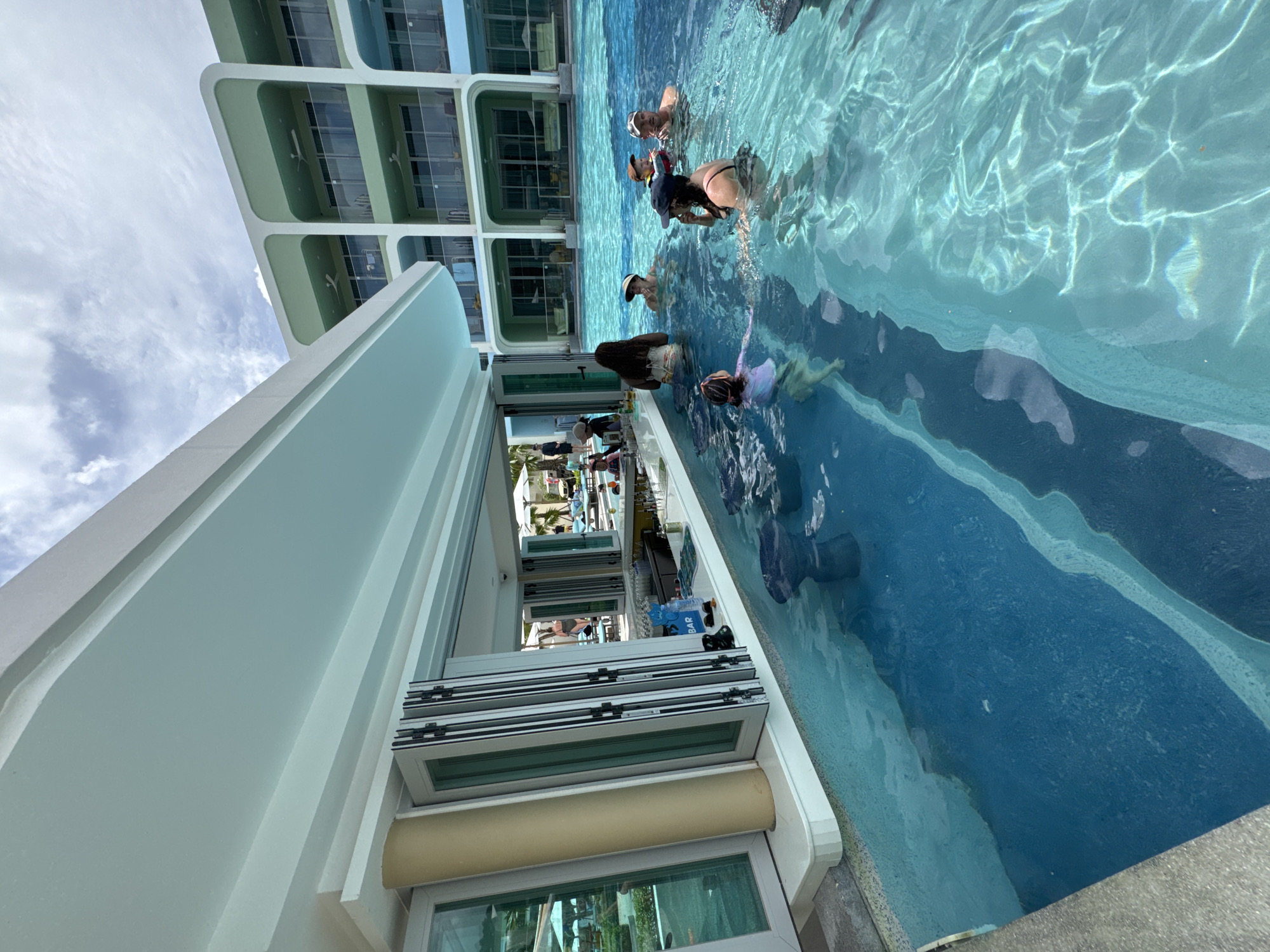 The image shows a scene in Karon, Thailand, likely at a resort or hotel. There is a multi-story building with glass balconies on the left side of the frame. Next to the building is a bar area with an open window-like structure that retracts upwards. People are in a swimming pool. Some are standing near the edge by the bar, while others are swimming further out. The pool water is clear and reflects the sky and buildings. In the background, one can see palm trees and other resort features through the opening in the bar area.