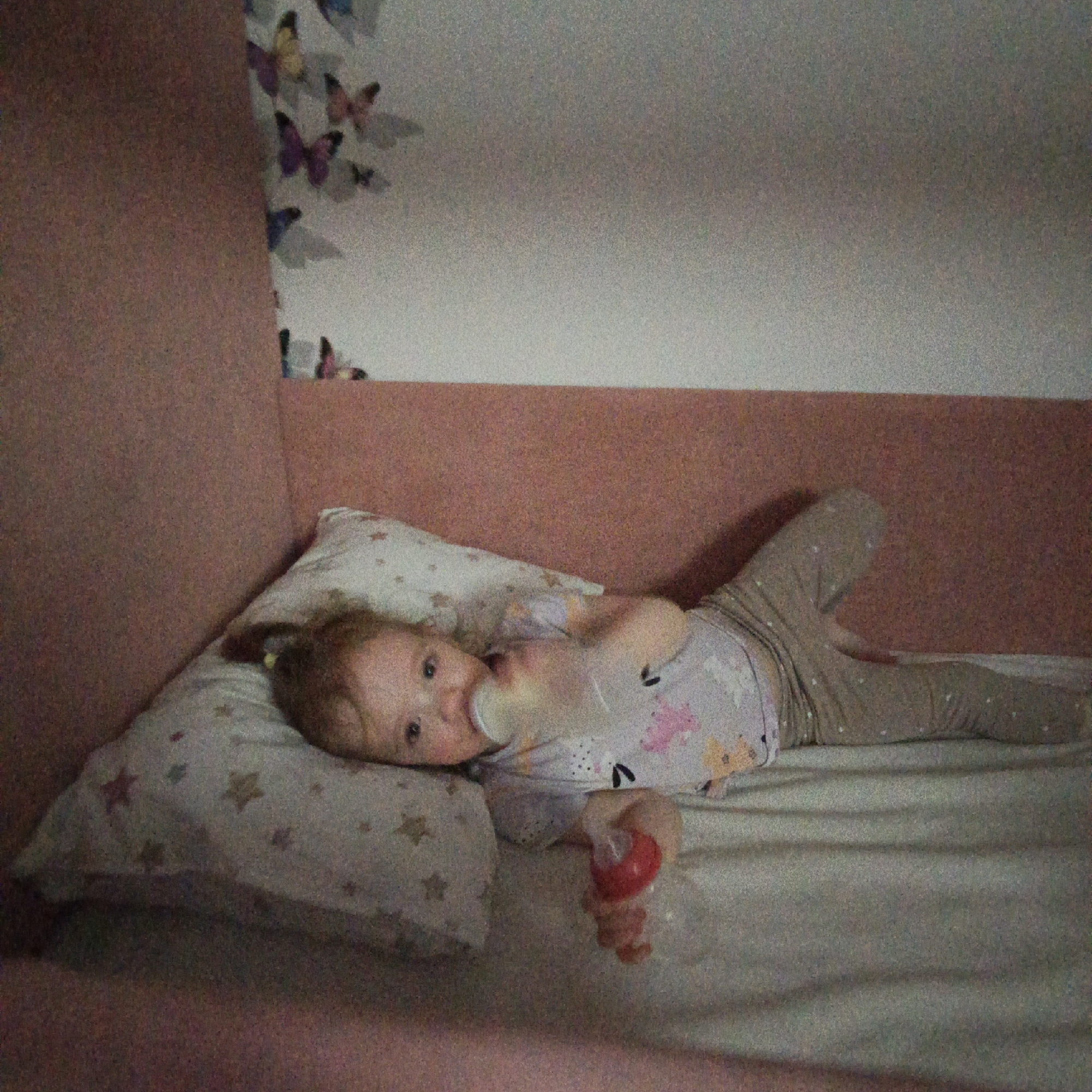 A small child is lying on a bed, seemingly in a bedroom. The child is wearing patterned pajamas and holding a baby bottle in their mouth. The background features a headboard and a wall decorated with butterfly stickers. The location is Марковский, Russia.
