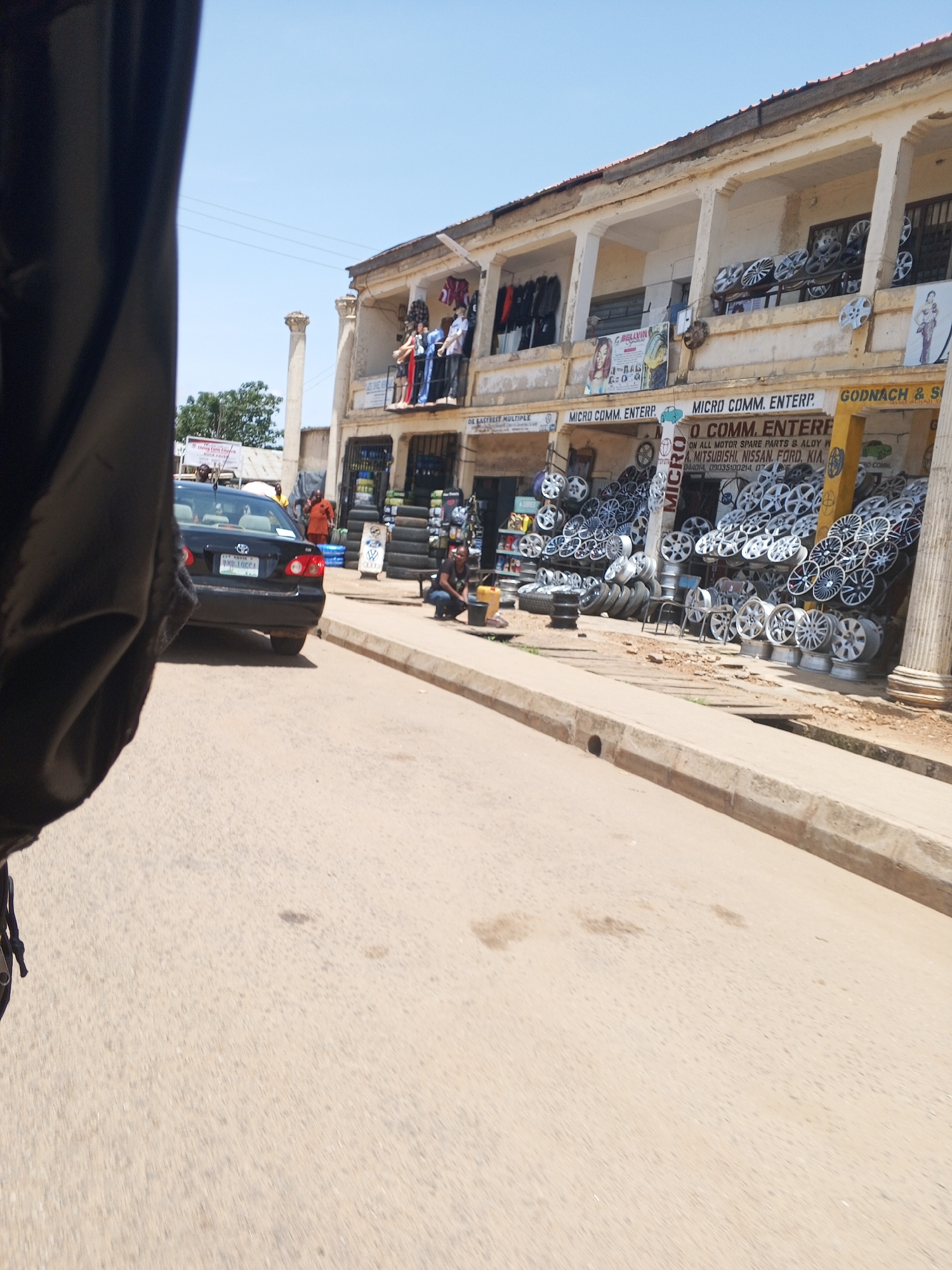 The image depicts a street scene in New Stadium, Nigeria, with a focus on a storefront selling car parts. The storefront is part of a multi-story building with a covered walkway supported by pillars. The walkway is lined with various car wheels, stacked and displayed for sale. Some tires are also visible, piled up in front of the store. A black sedan is parked on the road in front of the building. Two individuals are visible in the scene: one is a person squatting near the storefront, possibly attending to something on the ground, and another person in an orange garment stands near the car. The environment appears to be an urban street with a clear sky overhead. The image is taken from a slightly elevated perspective, likely from a moving vehicle.