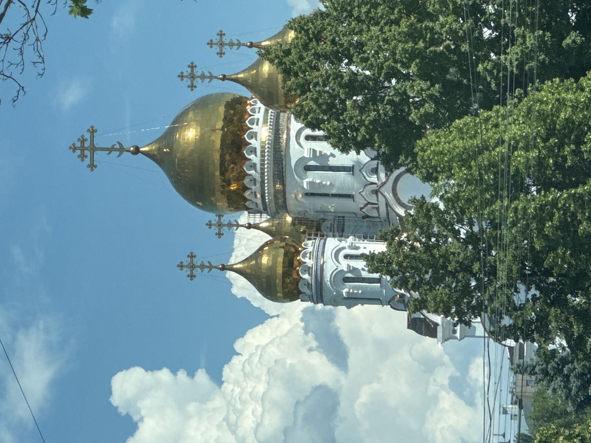 A white Orthodox church with gilded domes is partially obscured by green trees. The church has multiple onion-shaped domes topped with ornate crosses, characteristic of Orthodox architecture. The sky is blue with fluffy white clouds. Power lines are visible in the foreground, crossing horizontally across the lower part of the image. The scene appears to be on a sunny day.