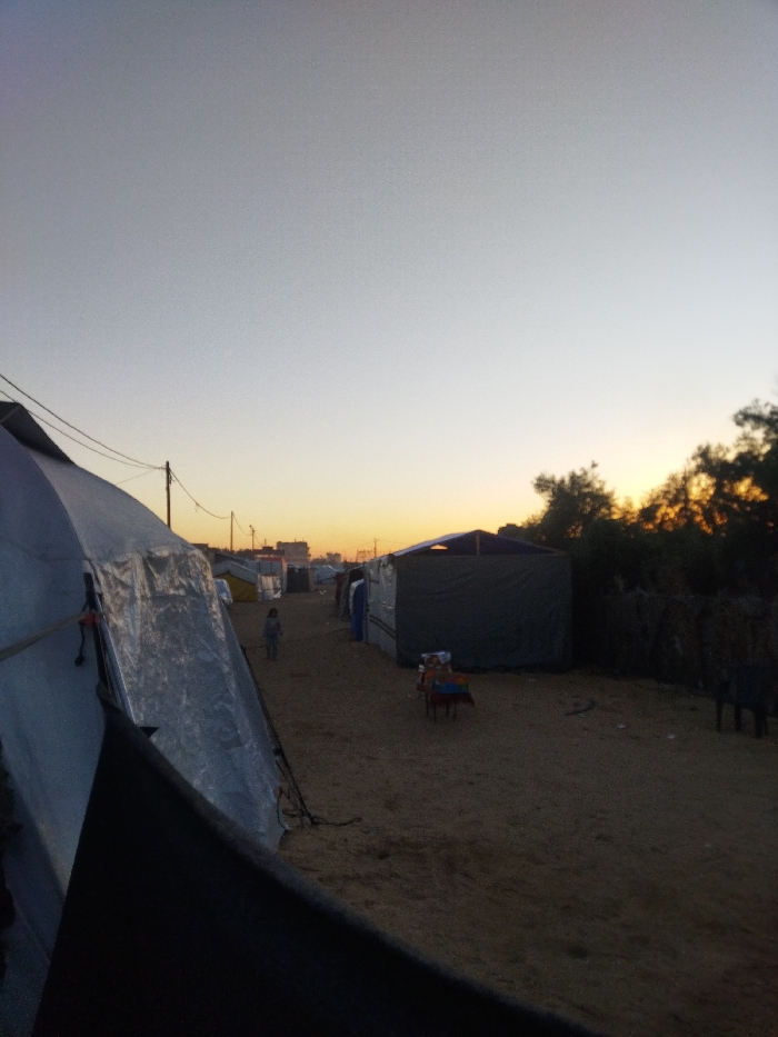 The image depicts an outdoor encampment in Khan Yunis, Palestinian Territory, at dusk. The sky transitions from light grey overhead to an orange and yellow gradient at the horizon. A wide, unpaved dirt path occupies the center of the frame, receding into the distance.
On the left, a large, light-colored tent or makeshift shelter, constructed from tarp-like material, dominates the foreground, secured with visible ropes and ties. Behind it, utility poles with electrical wires run along the left side of the path, diminishing in scale towards the horizon. In the mid-ground, a small child, dressed in light-colored clothing, stands alone on the dirt path, facing away from the camera.
On the right, a dark, rectangular, makeshift shelter with a lighter-colored, angled roof is visible. Adjacent to this shelter, a small, multi-colored chair or stool holds unidentified items. Further right, a dense line of trees or bushes, silhouetted against the setting sun, borders the encampment. Other indistinct structures and tents line the path in the background. The ground is composed of uneven, light-brown earth.