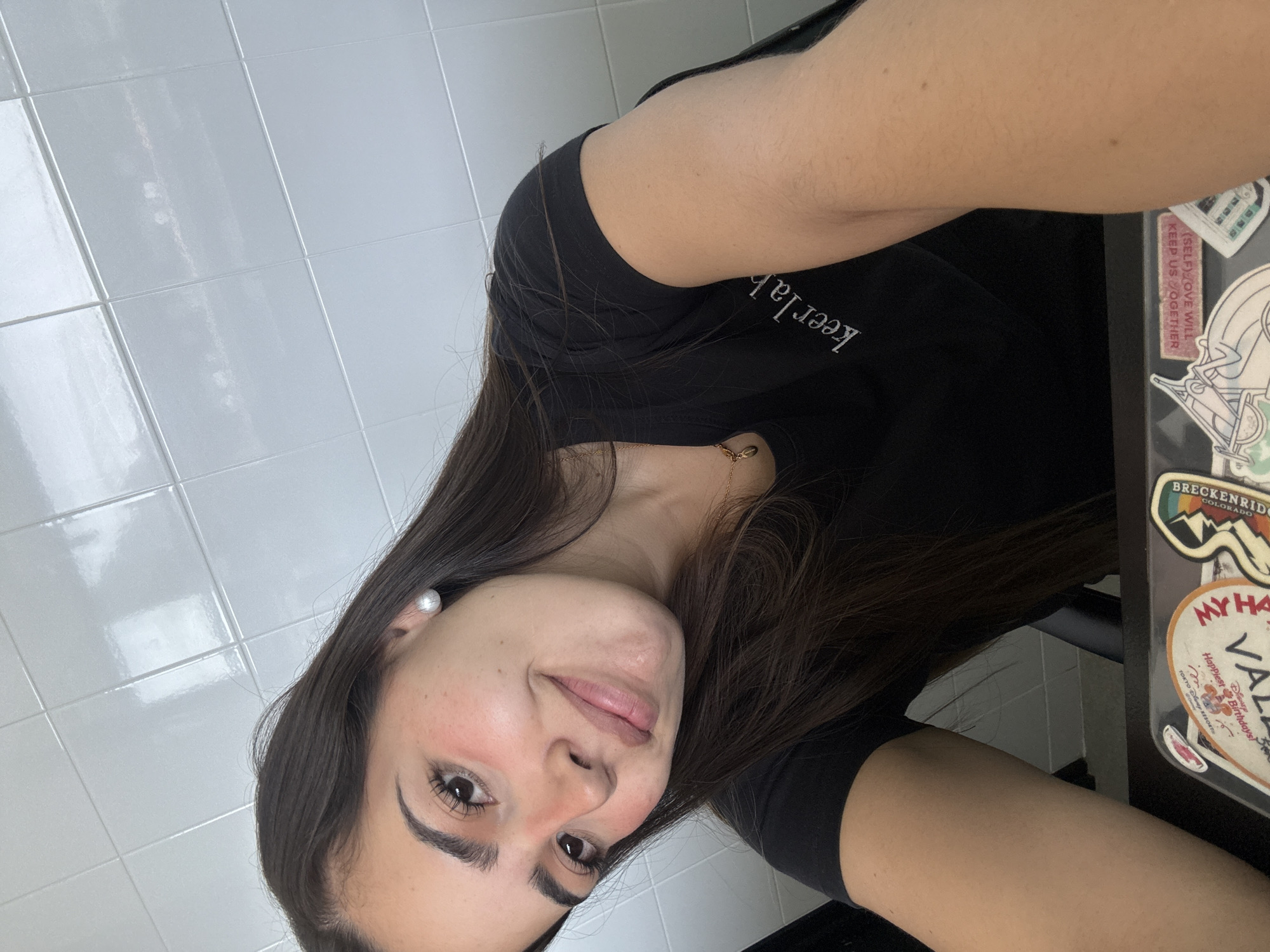 The image is a rotated, close-up selfie featuring a young woman with long, dark brown hair. She looks directly at the camera with a gentle smile and a pleasant expression; a noticeable reddish blush is present on her cheeks. She wears a black t-shirt with the white text "keerlab" visible on the shoulder. A delicate gold chain necklace is visible around her neck, and a pearl stud earring adorns her visible left ear.
The background to her left shows a bright white tiled wall with distinct grout lines, indicating an indoor setting, possibly a bathroom or a clean, domestic space. To her right, a portion of a dark laptop or notebook is visible, covered with various stickers.
Visible text on these stickers includes messages and place names: "SELF LOVE WILL KEEP US TOGETHER", "BRECKENRIDGE COLORADO", "MY HAPPY PLACE", "VALLE", and "Disney Happiest Birthdays TOKYO DISNEY RESORT". These details hint at her interests in travel and personal affirmations. The lighting appears even and diffused, consistent with an indoor environment. The context provided indicates the location is San Bernardino Tlaxcalancingo, Mexico, although no specific visual cues within the image confirm this.