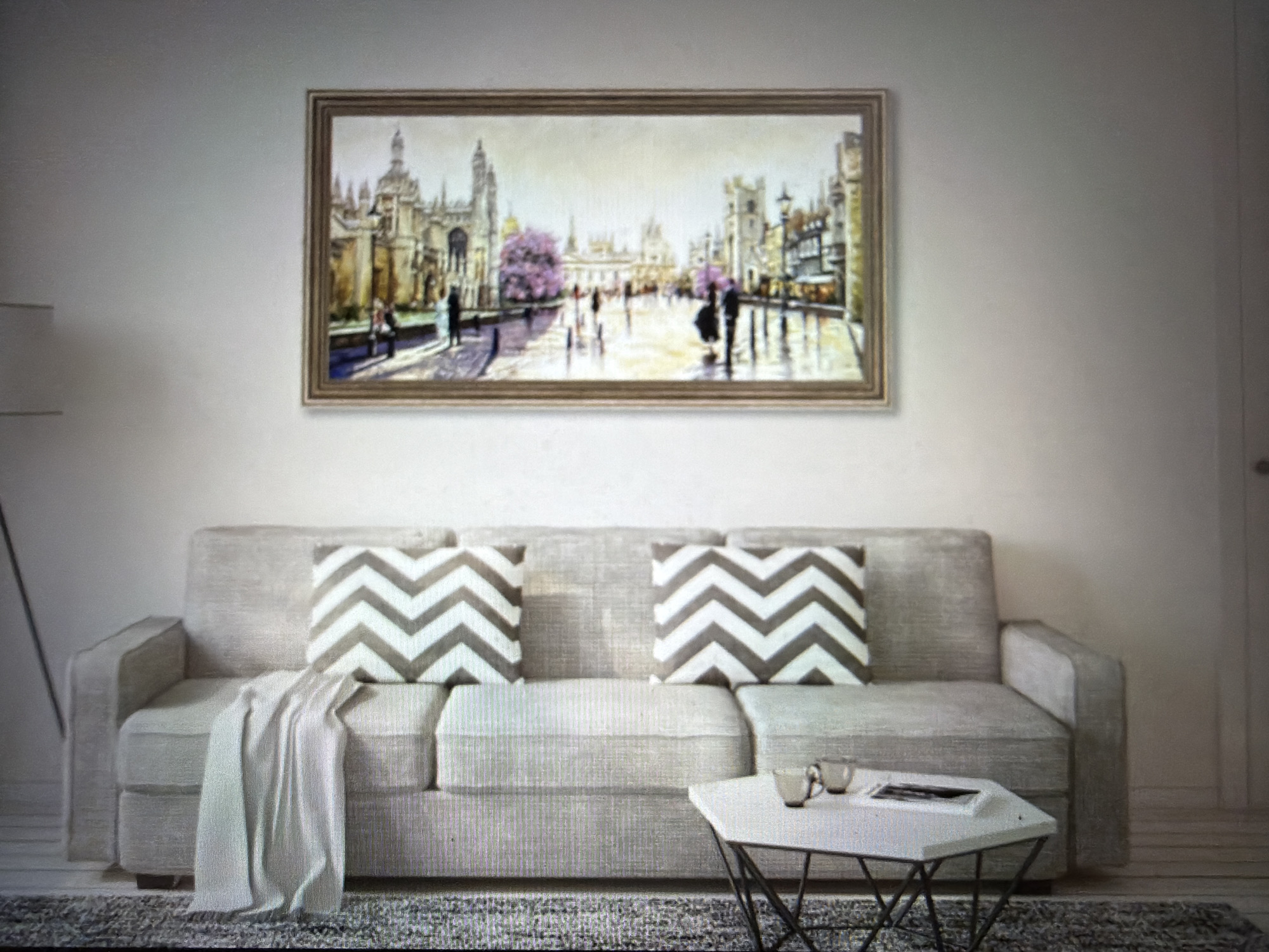 A horizontal framed painting depicting a city street scene with people and buildings is displayed on a light-colored wall above a light-colored sofa. The painting shows a wide street with ornate architecture on either side, and several figures in silhouette. A tree with pink blossoms is prominent in the mid-ground. The sofa has two chevron-patterned throw pillows and a light-colored throw blanket draped over one arm. In front of the sofa, a white, geometric coffee table holds two cups and a magazine. A floor lamp is visible to the left of the sofa. The overall setting appears to be a living room.