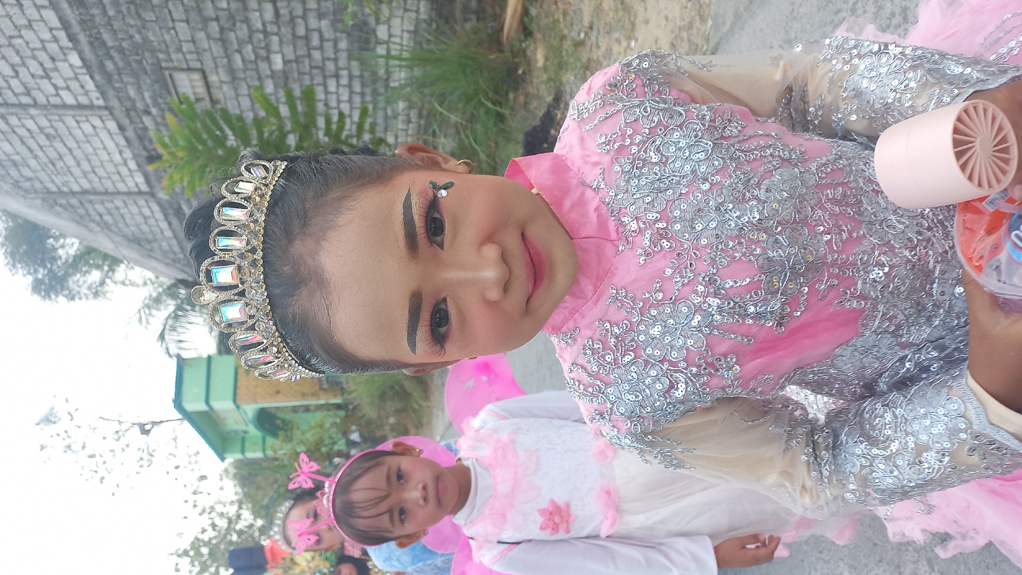 Two young girls, adorned in elaborate pink and silver sequined dresses and tiaras, are positioned in the foreground and middle ground of the image. The girl in the foreground, closer to the camera, wears a sophisticated tiara with large, colorful faux gemstones and a sequined pink and beige dress with a high neckline. She is looking directly at the camera with a gentle smile, her face professionally made up with dramatic eye makeup and rosy cheeks. She is holding a small pink fan in her right hand. The second girl, in the middle ground and slightly to the left, is wearing a simpler pink and white dress with lace details and a butterfly-themed headband. She is looking towards the camera with a neutral expression. Both girls appear to be participating in a parade or performance, as suggested by their attire.
The background reveals a blurred scene of other participants and an outdoor environment. To the left, a green and yellow arched gateway can be seen, possibly an entrance to a public space or school. A brick wall forms the right boundary of the scene. The ground is unpaved, suggesting a road or path. The overall setting appears to be an outdoor event in Kedungmegarih, Indonesia, given the context. The lighting is natural, indicating it is daytime, likely overcast or diffused sunlight.