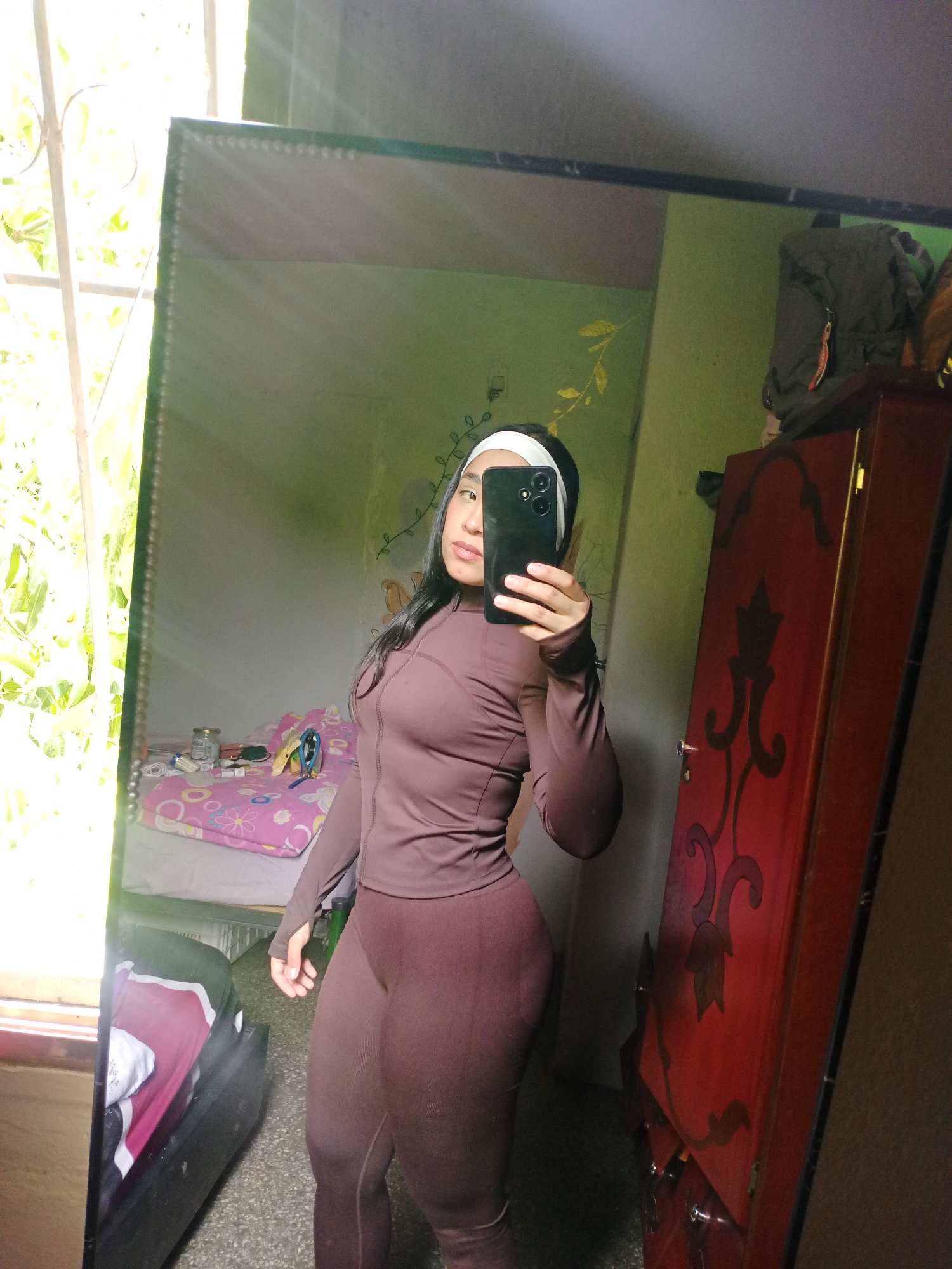 A young woman stands in a bedroom, taking a selfie in a mirror. She wears a fitted brown athletic suit and a white headband. The room contains a bed with a pink patterned comforter, a wooden wardrobe with a decorative design, and a window with greenery visible outside. A backpack sits atop the wardrobe. The location is likely a residential space within Valle de La Pascua, Venezuela.