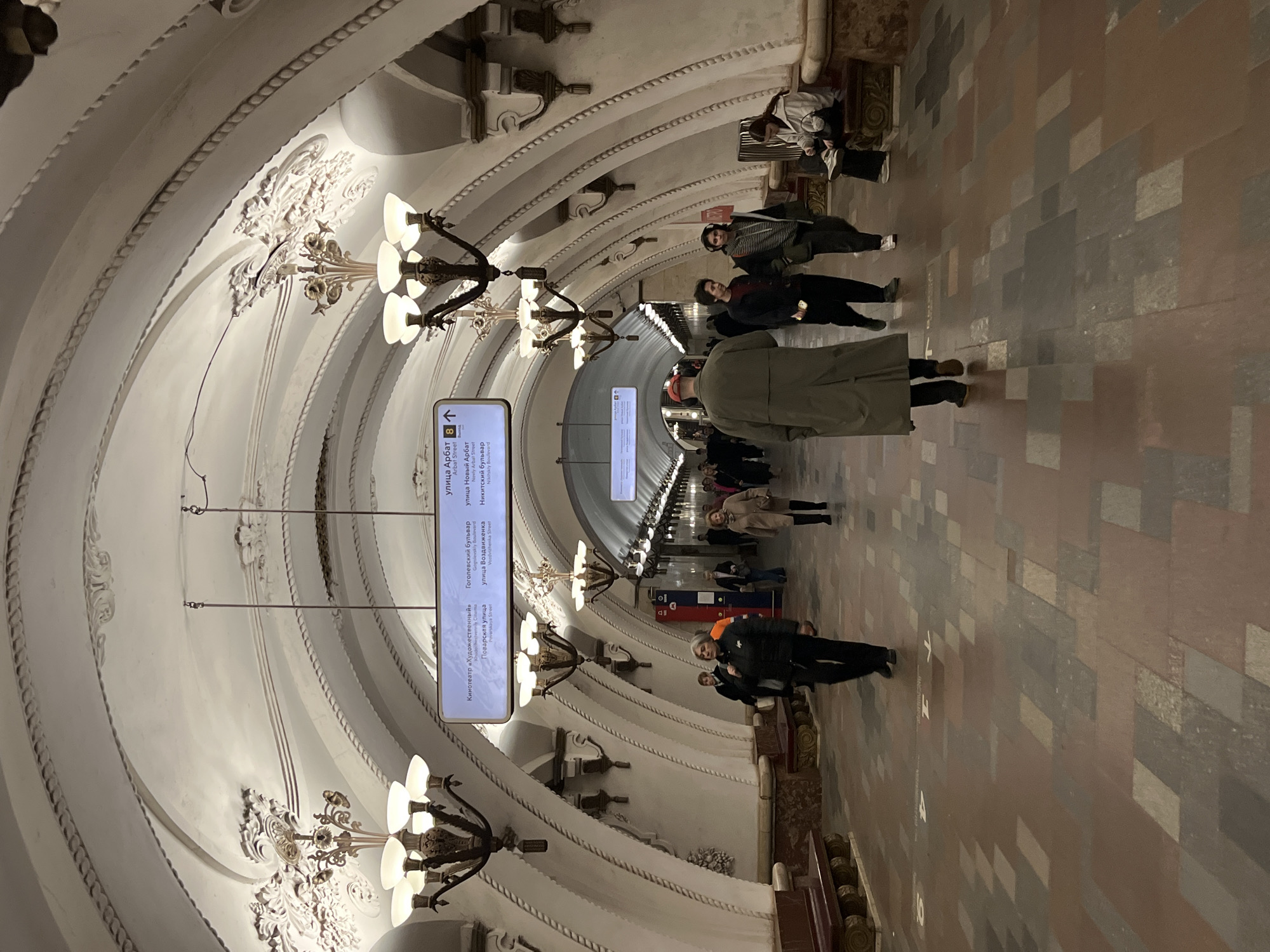 The image depicts an interior view of a metro station in Moscow, Russia. The station features a vaulted ceiling adorned with ornate plasterwork and multiple chandeliers. A sign displays information about local landmarks, including the Cinema "Khudozhestvenny," Povarskaya Street, Gogolevsky Boulevard, Vozdvizhenka Street, Arbat Street, Novy Arbat Street, and Nikitsky Boulevard. Several people are walking through the station. The floor is tiled in a pattern of light and dark rectangles.