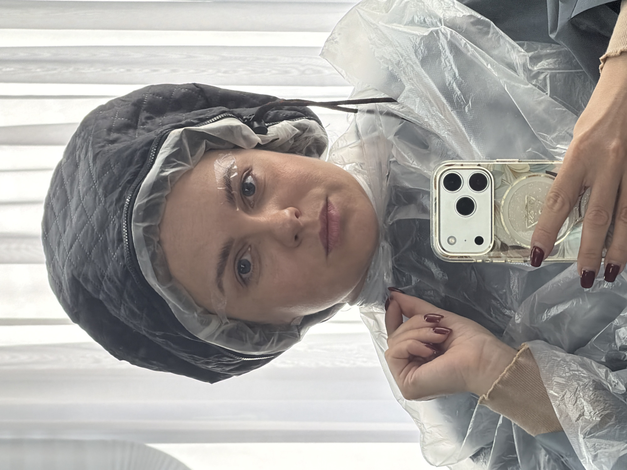 A woman is seen in a close-up selfie, wearing a black quilted cap over a transparent plastic hair covering. Adhesive strips are visible on her eyebrows. She holds a smartphone with a clear case and a decorative circular pop socket. Her nails are painted a dark red color. The background features horizontal white blinds. The image does not provide enough information to determine the location as Votkinsk, Russia.
