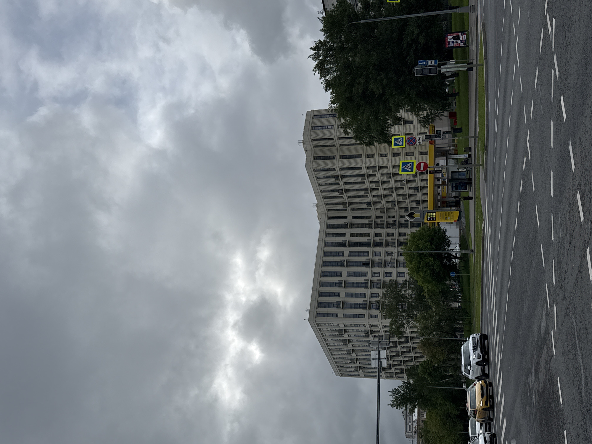 The image depicts a city street scene under an overcast sky. On the right, a paved road stretches into the distance, marked with white dashed lines indicating lanes. A line of vehicles, including cars and trucks, are parked or moving along the roadside. Adjacent to the road, a green strip of grass and trees separates the street from a tall, multi-story building. The building is characterized by its repetitive window pattern and modern design. In front of the building, various street signs and possibly a gas station are visible. The sky is predominantly gray with dense clouds, suggesting a cloudy or rainy day. The photograph appears to be taken at ground level, offering a street-level perspective of the urban environment.