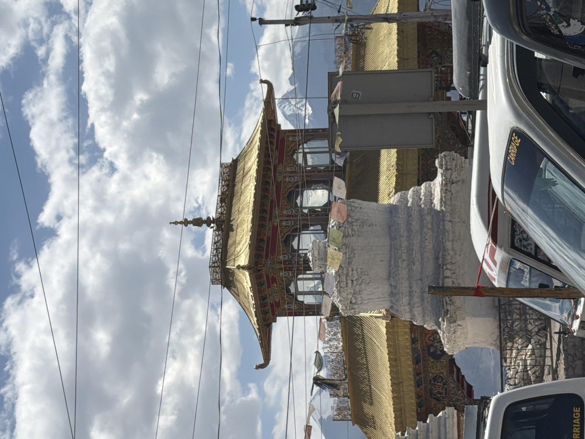 The image shows a view of a Buddhist monastery with golden roofs, white stupas, and prayer flags in the city of Leh, India. The monastery's architecture is ornate, with red and gold accents and intricate carvings. In the foreground, several vehicles, including a white van with the word "CARRIAGE" on its windshield, are parked. In the background, snow-capped mountains are visible under a cloudy sky. The scene appears to be a street in Leh, a city known for its Buddhist heritage and proximity to the Himalayas.