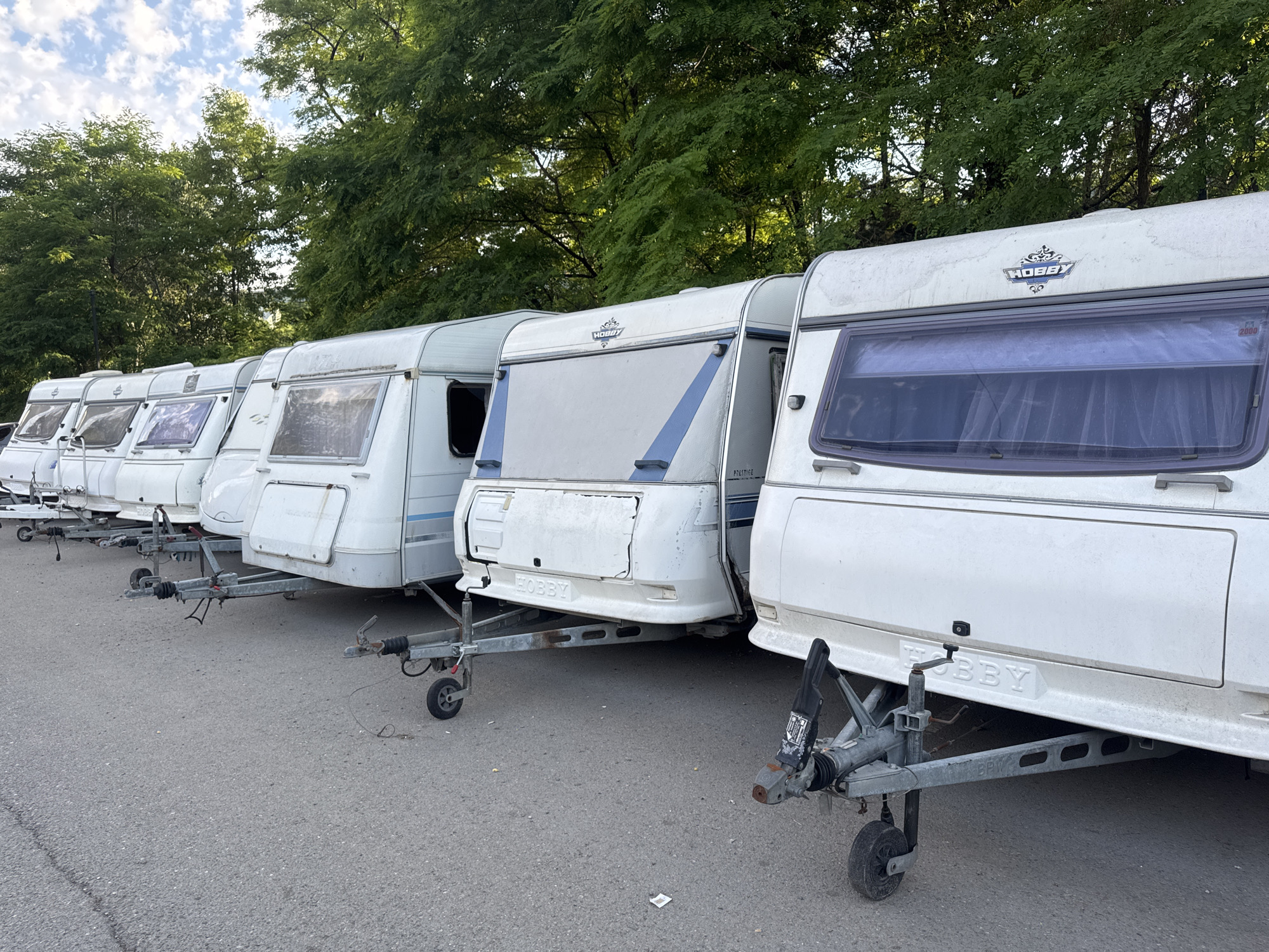 The image shows a row of white Hobby brand caravans parked on an asphalt surface. The caravans are lined up side-by-side, with trees and foliage visible in the background. The sky is cloudy. The caravans appear to be in a storage or parking area. The scene is captured from a low angle, focusing on the lower parts of the caravans and their trailers. There are no people or animals visible in the image. The setting appears to be outdoors, in a natural environment with a man-made parking surface.