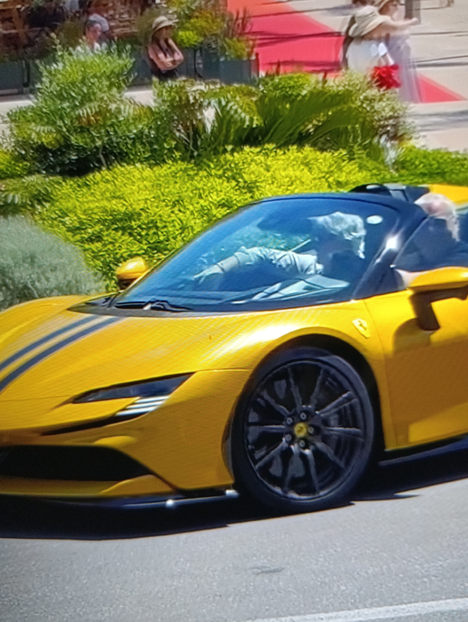 The image shows a yellow Ferrari SF90 Stradale parked on a paved road, likely in a public area, as indicated by the presence of people and greenery in the background. The car features black racing stripes on the hood, black wheels, and a black roof panel. The driver is visible inside the car. In the background, there is lush green vegetation, and indistinct figures of people walking on a red carpet or elevated platform. The location appears to be an urban or resort area with landscaping.