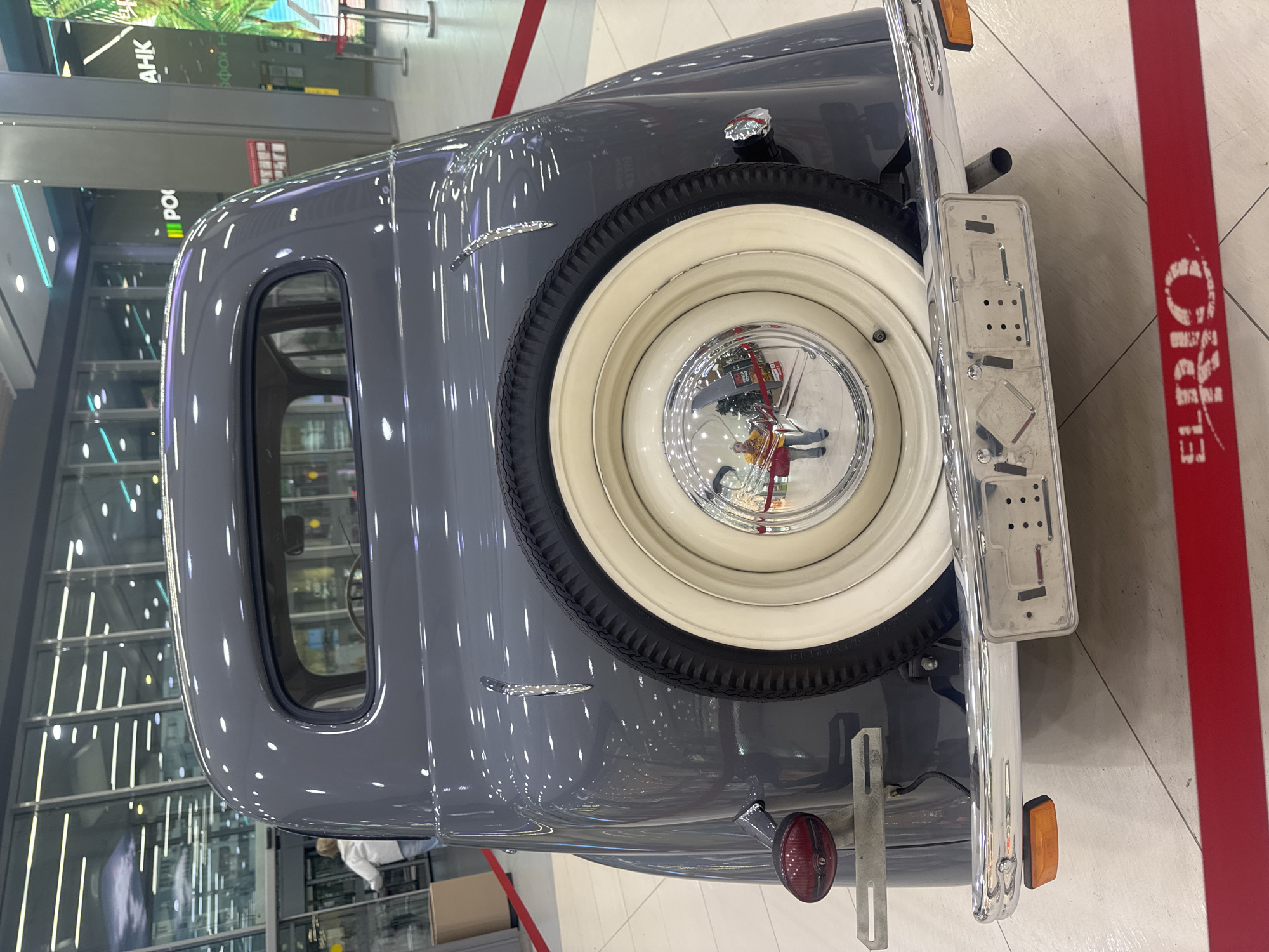 The image shows the rear of a vintage grey car parked indoors, likely in a shopping mall or exhibition space. The car has a spare tire mounted on the back with a white rim and chrome hubcap. The hubcap reflects the surrounding environment, showing people and a Christmas tree. A chrome license plate holder is also visible, attached to the rear bumper. The car has a small red taillight and an orange reflector. The setting appears to be a brightly lit indoor environment with polished floors and reflective surfaces. In the background, there are signs with Russian text, including "AHK" and "РОС". The overall impression is of a well-preserved classic car on display.