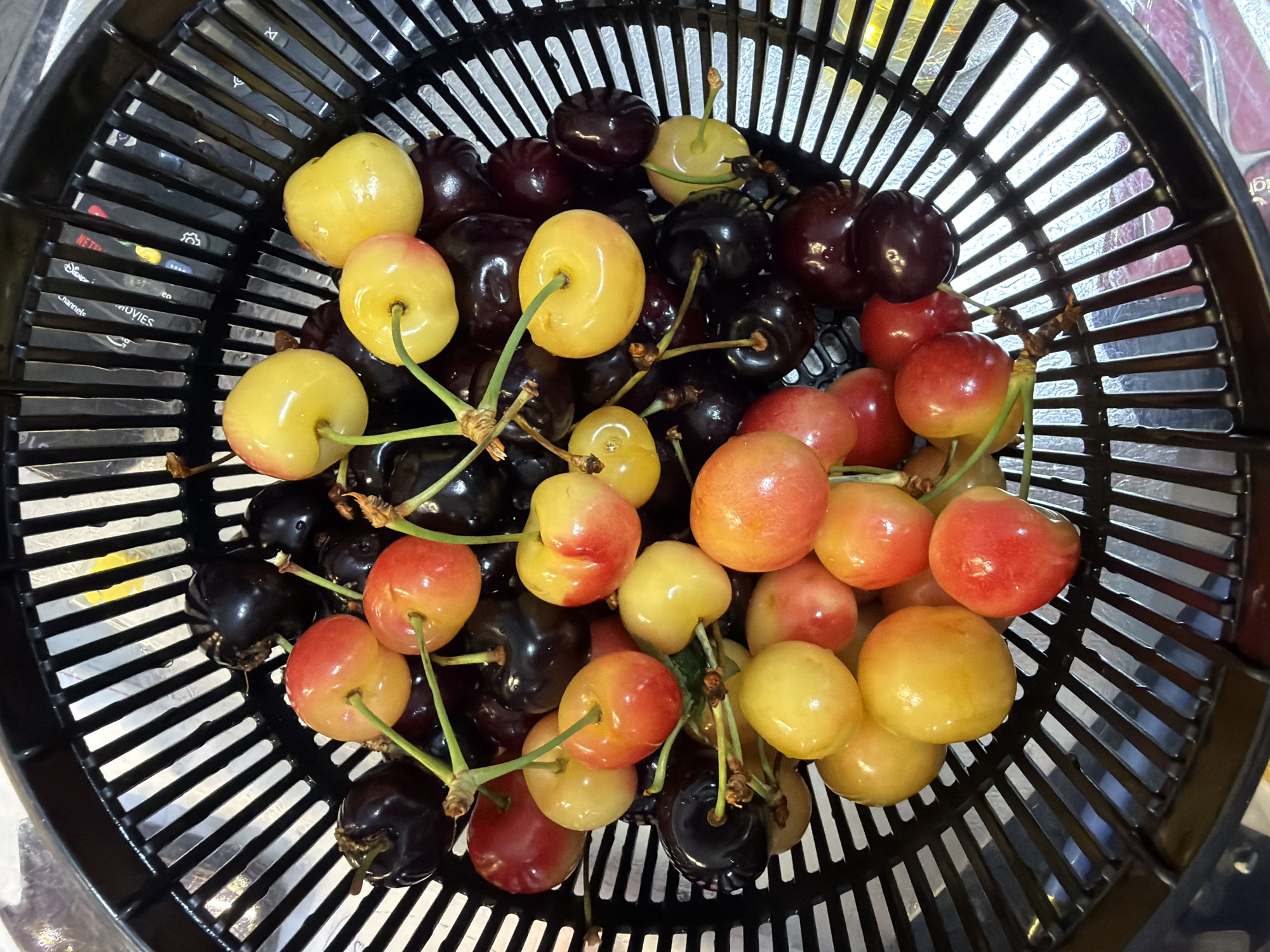A black plastic colander is filled with a mixture of Rainier and dark cherries. The cherries vary in color from bright yellow with red blush to deep red and dark purple. Some cherries have stems attached. The colander is positioned over a reflective surface, possibly a countertop or table, and the background shows a blurred interior setting with some handwritten notes on a white surface. The image does not provide any information that allows for the localization of this scene in Ashkelon, Israel, or any specific city.