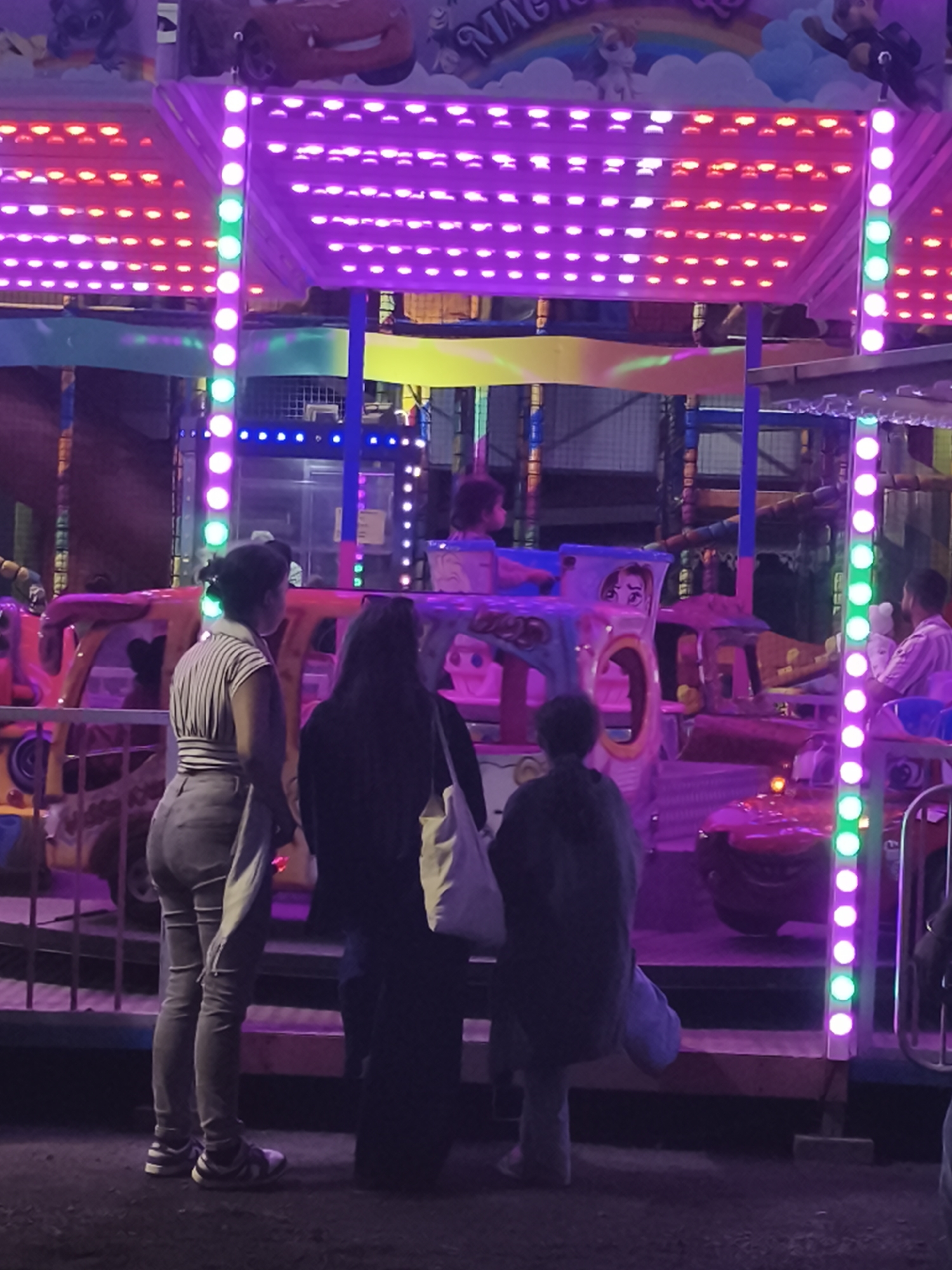 This image captures a scene at a nighttime amusement park or fair in Saint-Denis, France. The foreground features three individuals standing and observing. The person on the left, facing away from the camera, wears a striped shirt and light-colored pants. To their right, another individual, also facing away, is dressed in dark clothing and carries a white tote bag. The third person, on the far right, is only partially visible and appears to be wearing jeans.
Behind these individuals is a brightly lit amusement ride, adorned with numerous pink and red LED lights. The ride itself has colorful, cartoon-like elements, including a depiction resembling characters from the movie "Cars" on an overhead banner. People are visible on the ride, suggesting it is in operation. The ground is dark and appears to be a mix of dirt and gravel. The overall atmosphere is one of evening entertainment, characterized by the vibrant artificial lighting.