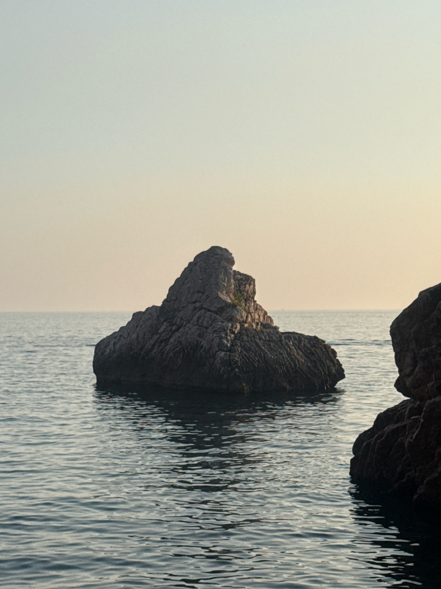 A solitary, large, dark rock formation emerges from the calm, rippling water of the Adriatic Sea. The rock is rough and textured, with visible fissures and crevices. A small patch of green vegetation grows from the top of the rock. The water is a deep blue-gray, reflecting the soft, hazy light of a late afternoon or early morning sky. The sky itself is a gradient of pale yellow and light blue, with no visible clouds. To the right of the frame, a portion of another large, dark rock, likely connected to the shore or a larger formation, is visible, its textured surface contrasting with the smooth expanse of the water. The overall scene evokes a sense of tranquility and natural isolation. This scene is located off the coast of Rafailovići, Montenegro, a coastal village known for its picturesque beaches and clear waters.