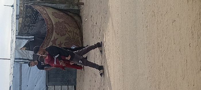 An outdoor scene in Khan Yunis, Palestinian Territory, depicts several individuals in a temporary settlement environment under a light overcast sky. In the foreground, a young adult male with short dark hair and a beard, wearing a black long-sleeved shirt, dark grey pants, and light-colored slides over dark socks, walks left-to-right across the frame. He carries a small child, dressed in red, held close to his left chest. The child's face is obscured. Behind them, to the left, another young male in a black t-shirt looks towards the left. Further right and partially obscured, a third individual in red is visible. The ground is unpaved, consisting of light-colored dirt or sand. In the background, makeshift shelters are visible: one to the left is constructed with light-colored tarpaulin and features a metal door; another to the right is covered with brown patterned blankets or tapestries, forming an opening.